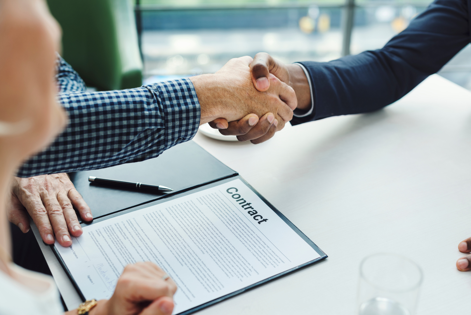 Two people shaking hands over a contract on a desk, with one person's hand visible and the other person's hand extended. A pen and a sheet of paper titled 'Contract' are also seen on the desk.