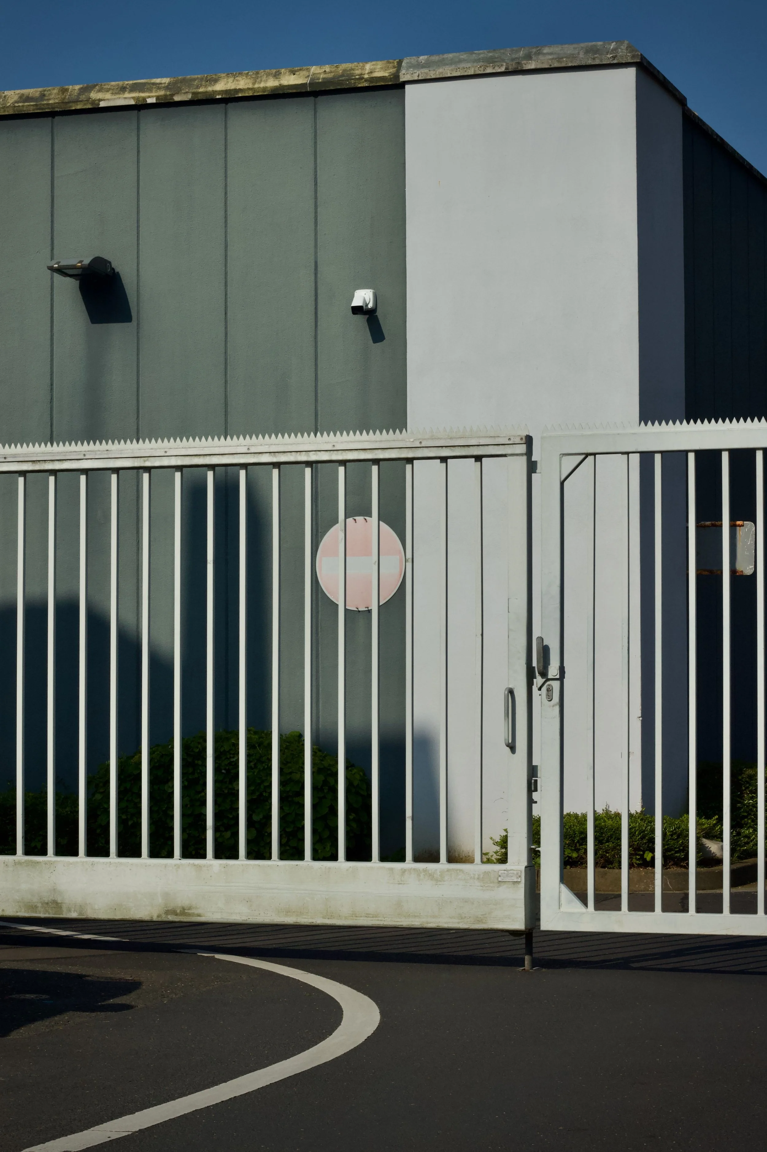 A building with a gray and white exterior wall and a white gate with a lock. There is a security camera and a vent on the building's wall. The scene is set outside on a clear day with a blue sky.