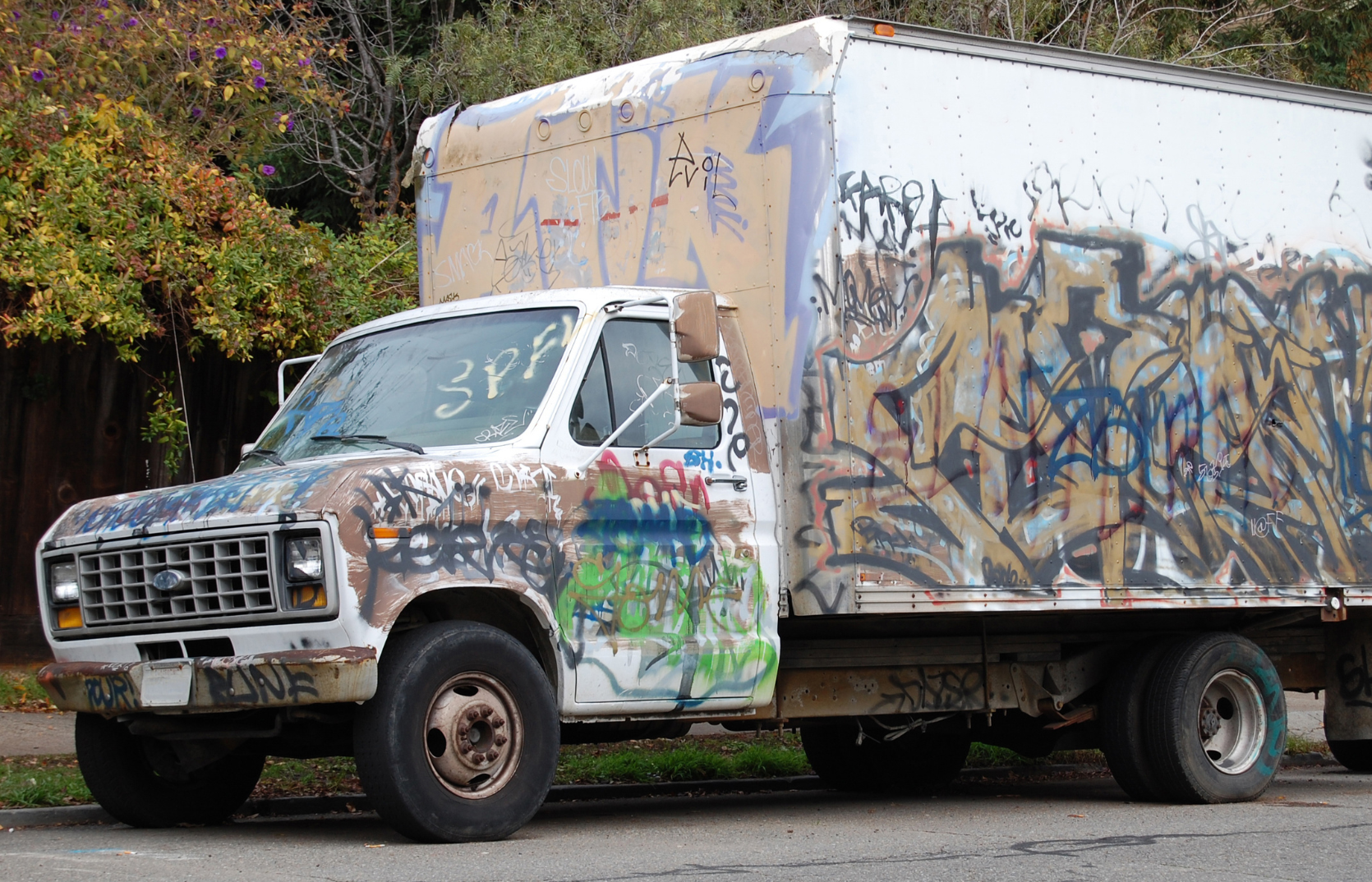 An old, vandalized, white delivery truck covered in colorful graffiti and spray paint, parked beside a sidewalk with green and purple foliage in the background.