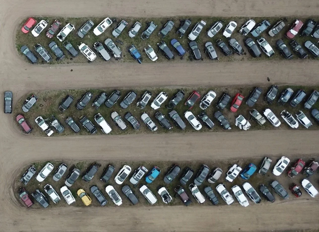An aerial view of a parking area with rows of parked cars on a dirt lot, separated by dirt paths, some parked on grassy patches, with a mix of colors and types of vehicles.
