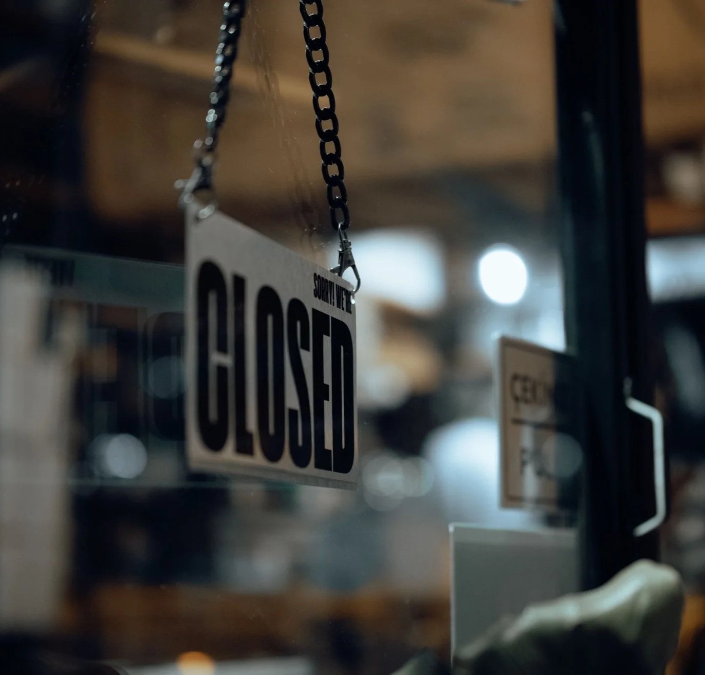 Close-up of a sign hanging from a chain that says 'Sorry We Are CLOSED' in bold black letters on a white background, with a blurred background and visible reflections in a window.