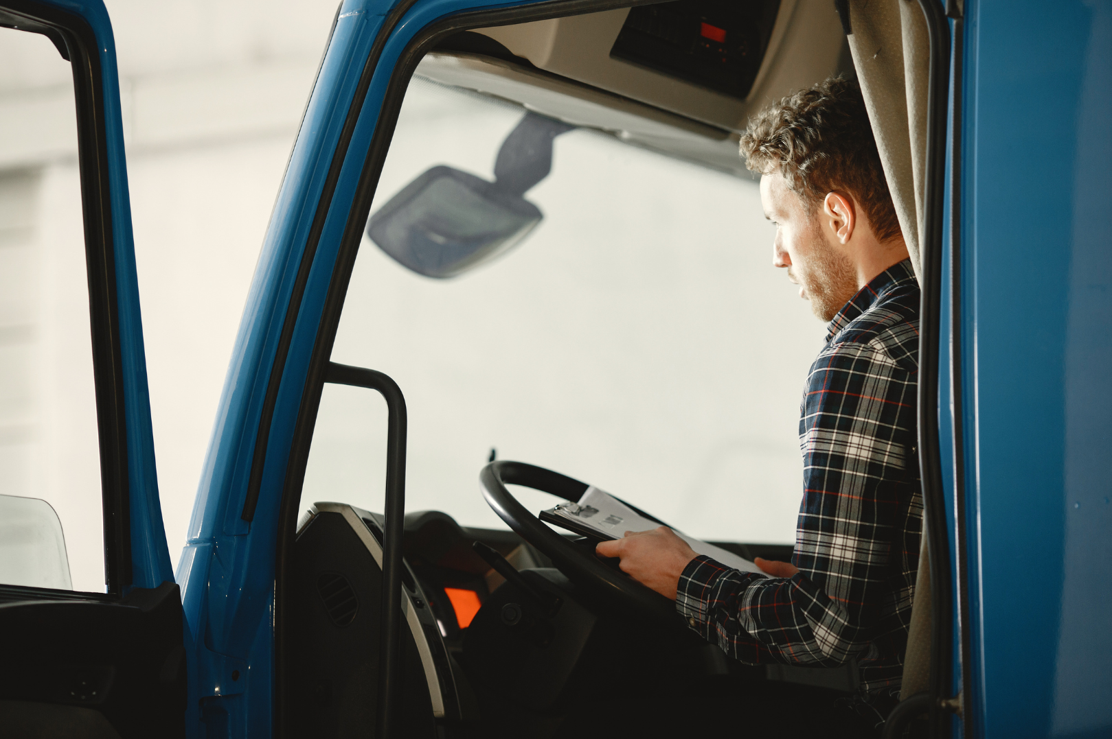 A man with curly hair and a plaid shirt sitting in the driver's seat of a blue semi-truck, looking at a tablet.