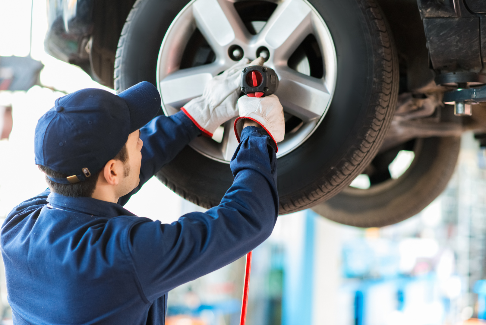 A mechanic working on a car tire in a garage, wearing a blue uniform and white gloves, with a screwdriver in hand.