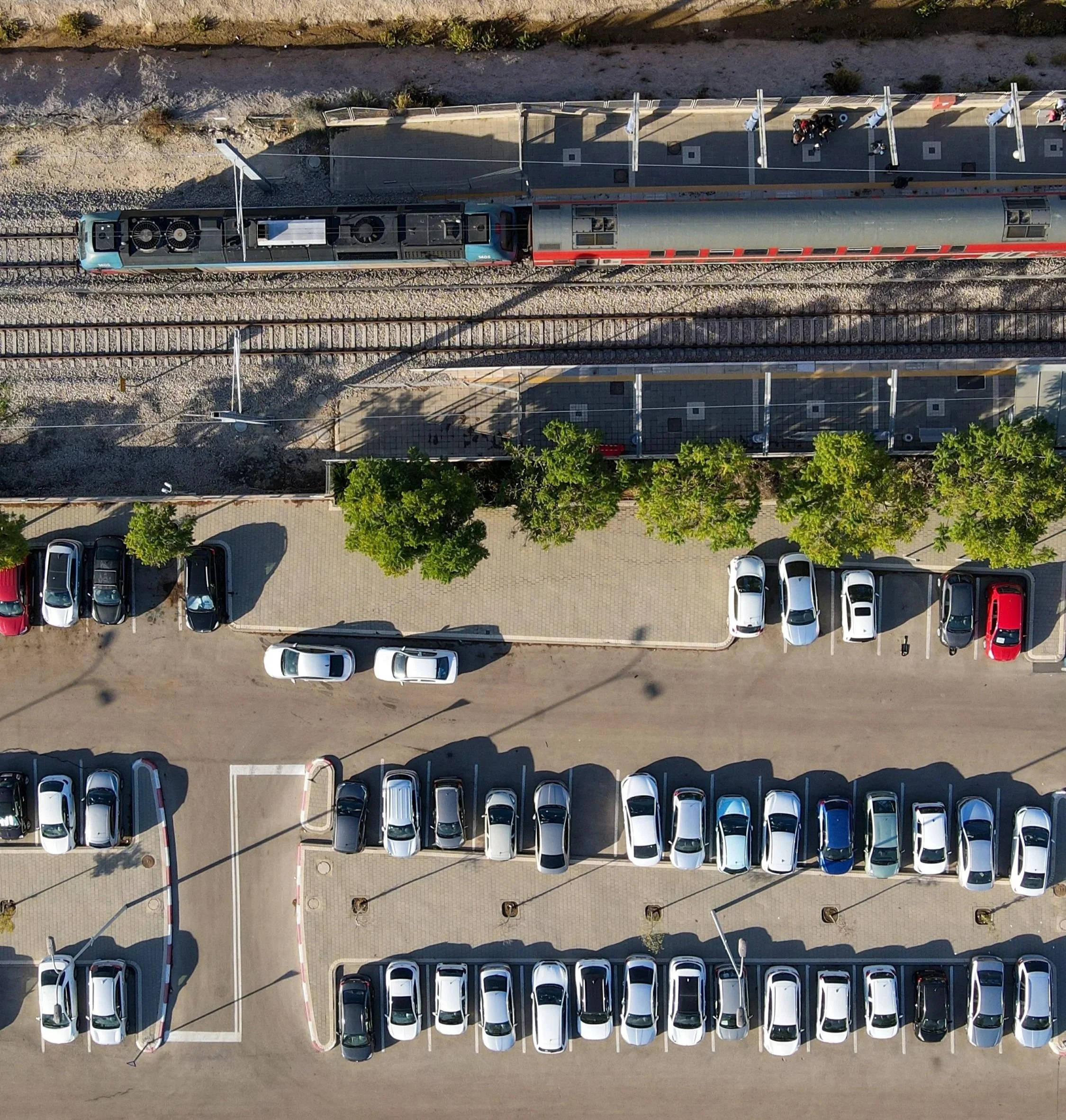 An aerial view of a parking lot with organized rows of white cars and trees, a train passing on railway tracks adjacent to the parking lot.