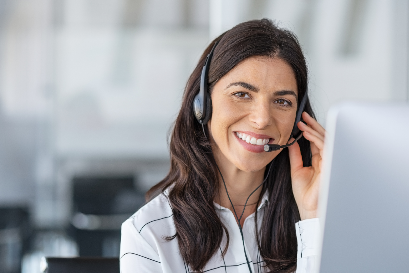 A smiling woman wearing a headset with a microphone, working at a computer in an office setting.