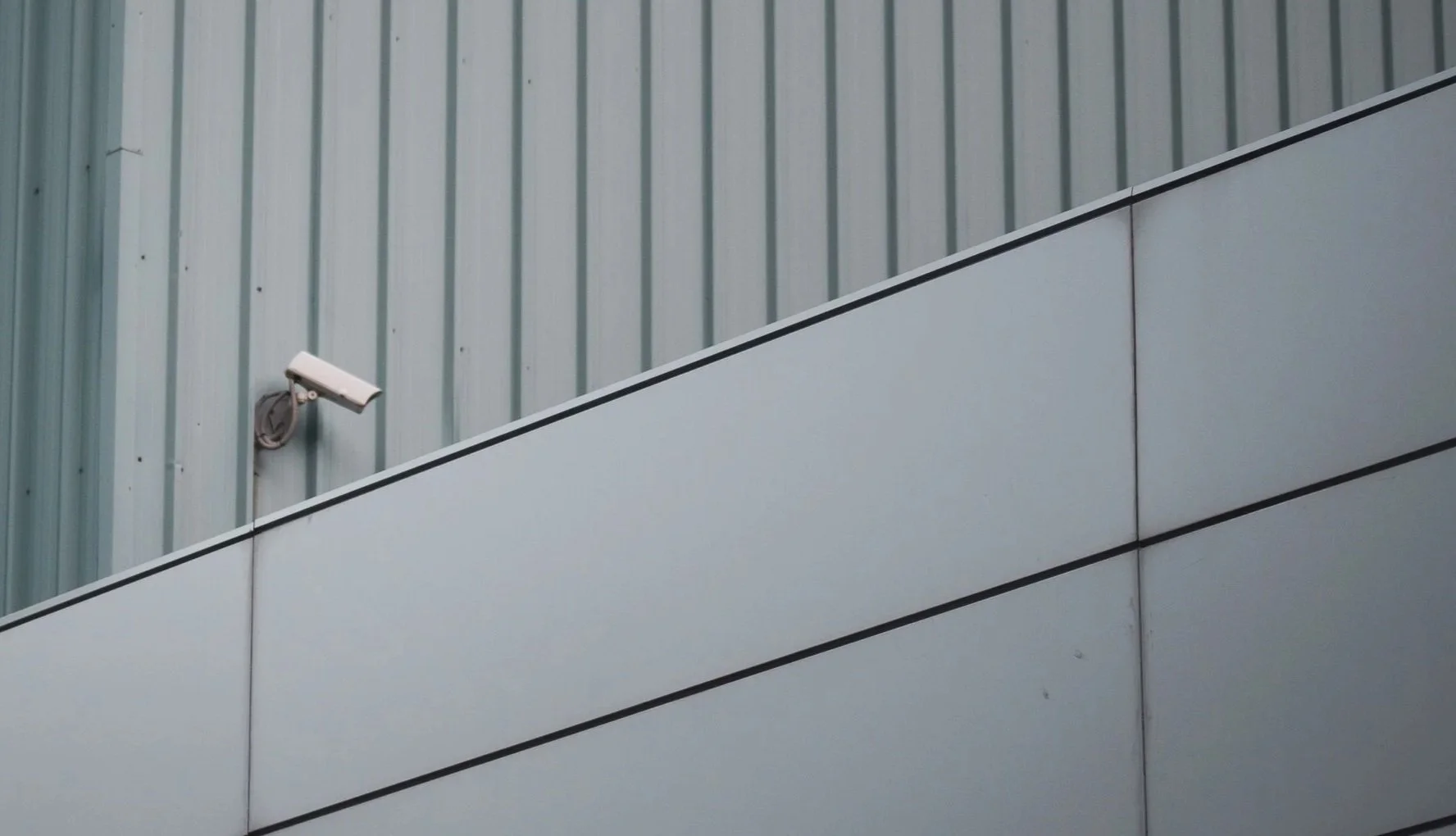 Close-up of a surveillance camera mounted on a building with metallic siding and a horizontal white panel, photographed from below.