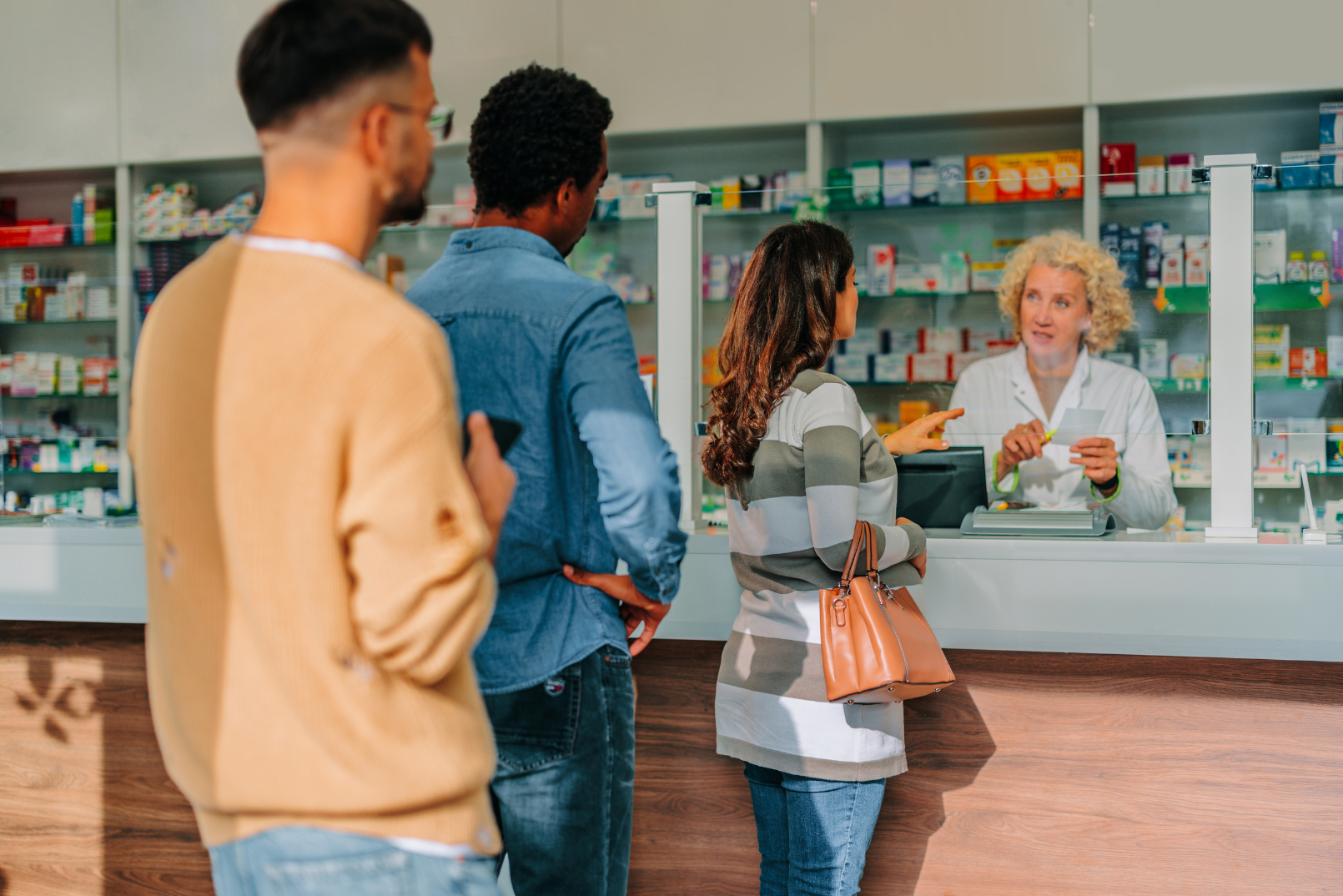 People standing at a pharmacy counter being assisted by a pharmacist. The pharmacist is showing a prescription to a woman who is holding a brown purse. Two other individuals are waiting in line behind her.