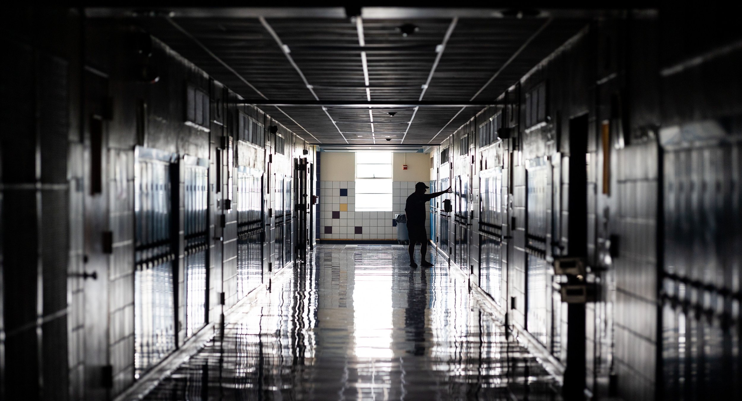 A person standing in a hallway of school lockers, reaching for an item or door handle, with natural light coming from a window at the end of the hallway.