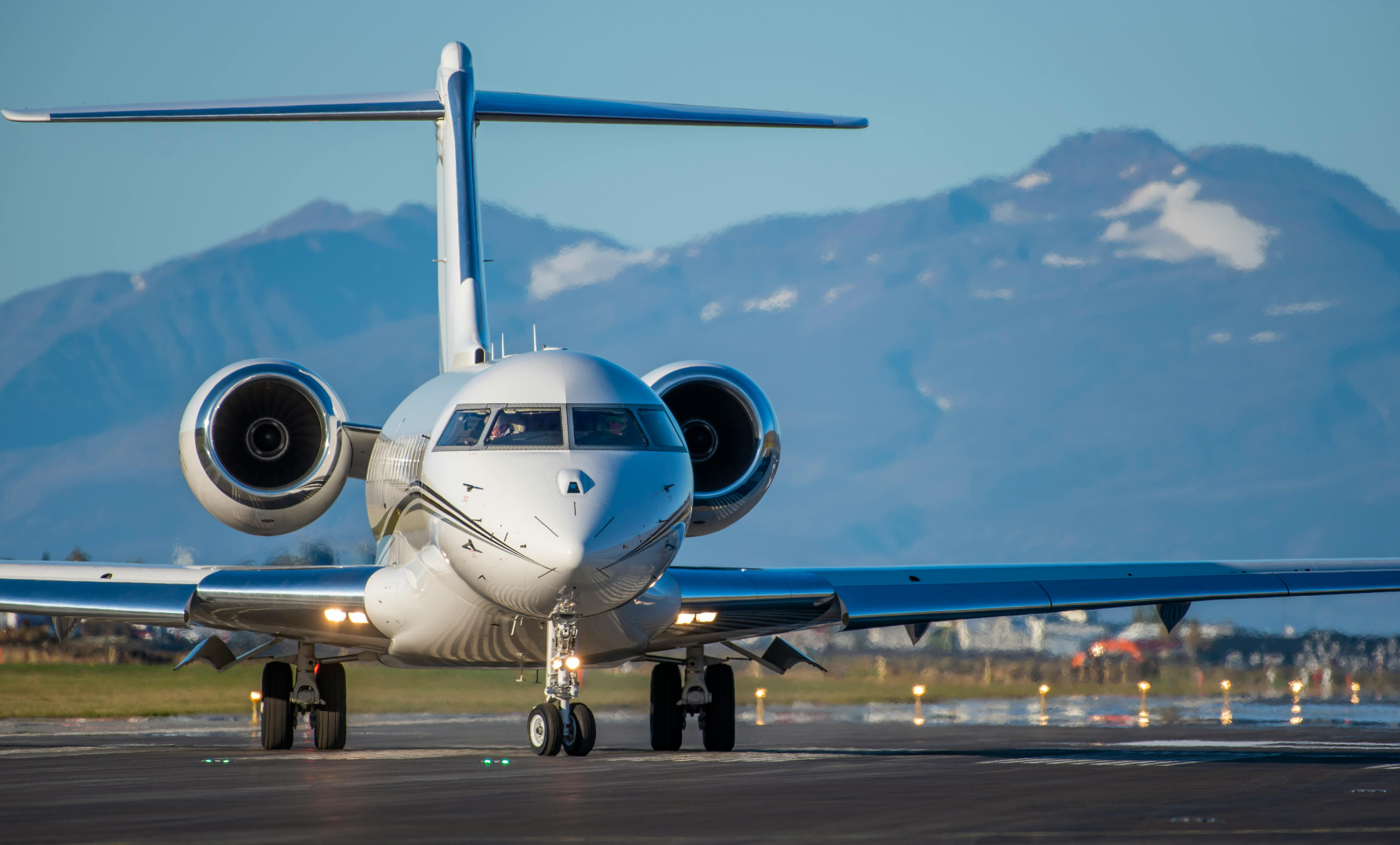 Front view of a white private jet on the runway with mountains in the background.