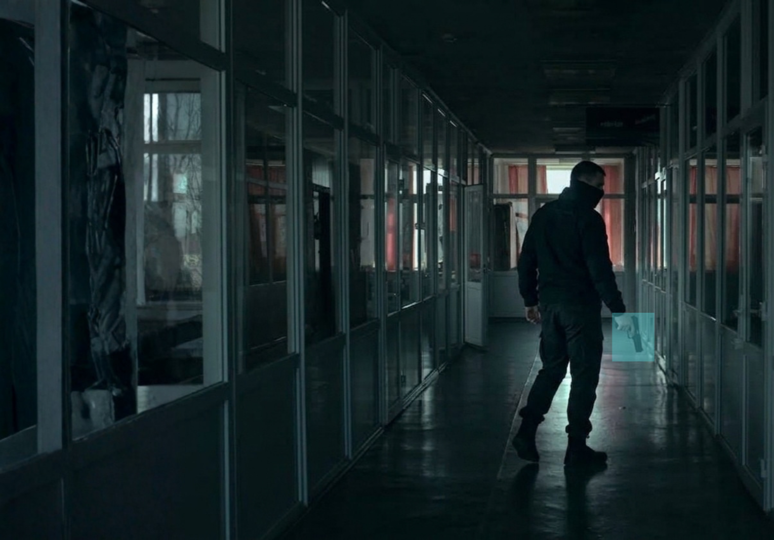 A person dressed in black with a face mask and sunglasses holding a gun in a dark, empty hallway.