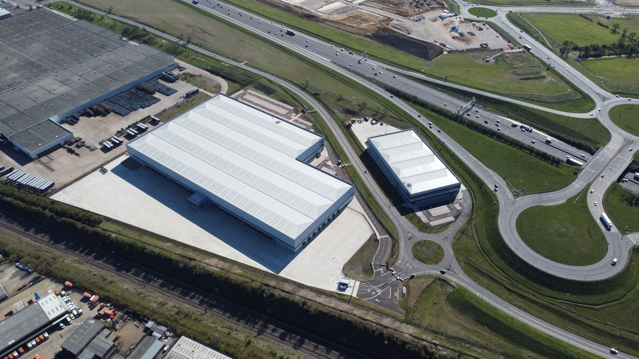 An aerial view of an industrial area with large white buildings, roads, a railway, and surrounding greenery.