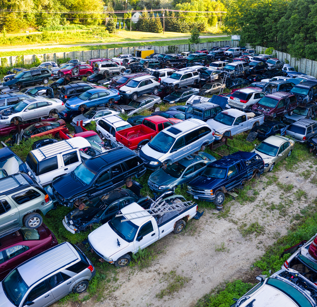 Aerial view of a junkyard filled with various abandoned and damaged cars, surrounded by trees and a wooden fence.