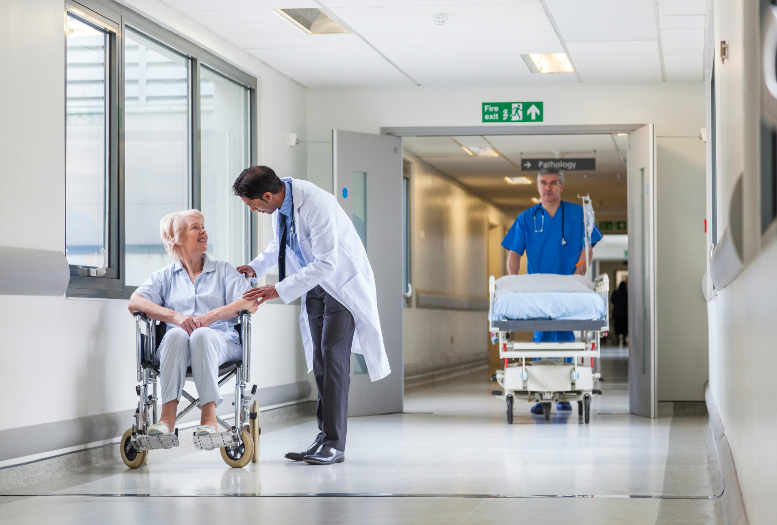 An elderly woman in a wheelchair being assisted by a doctor in a hospital corridor, with a nurse pushing a hospital bed in the background.