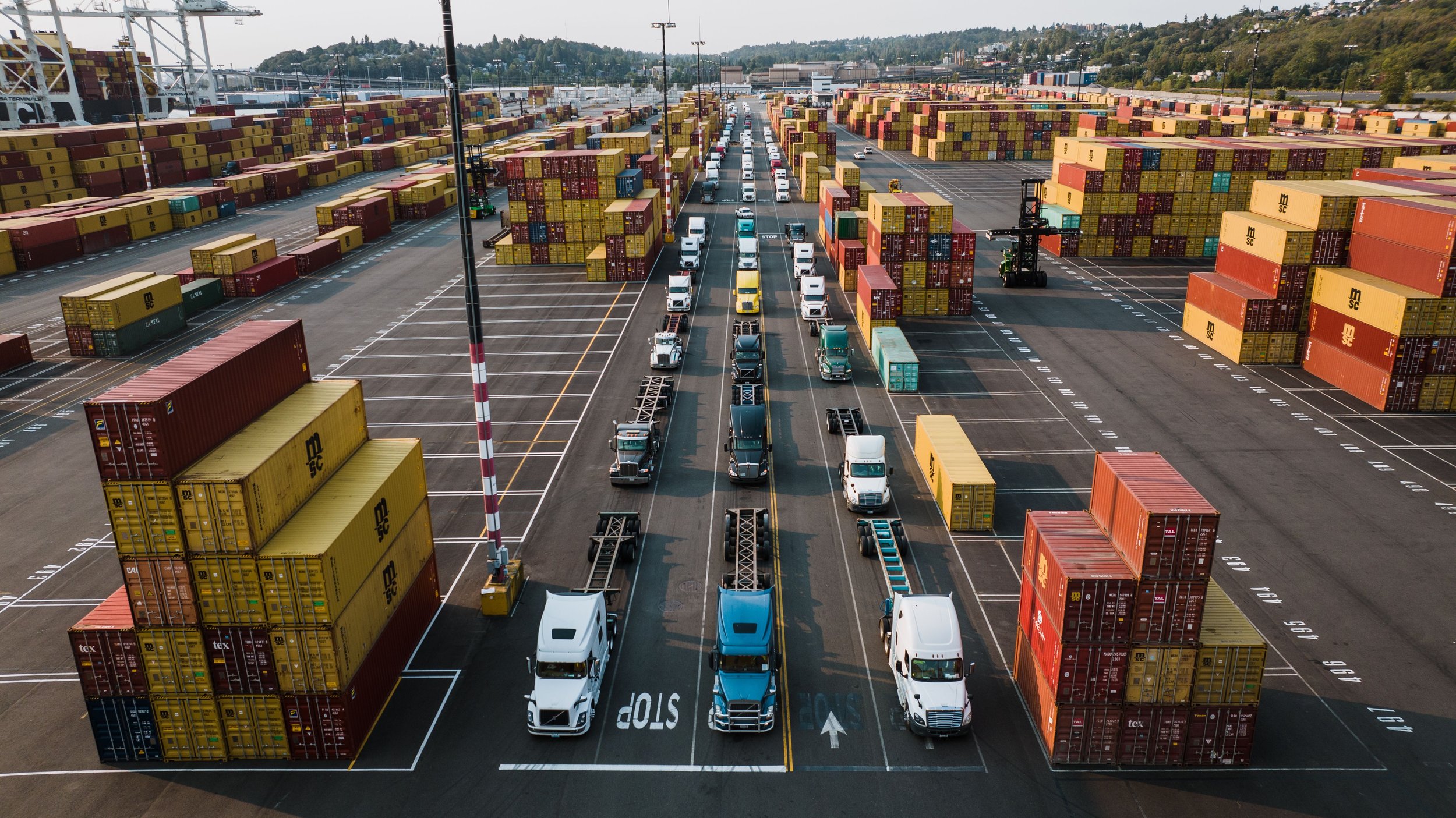 A large container port with numerous shipping containers stacked and trucks lined up for loading and unloading, with cranes and infrastructure visible in the background.