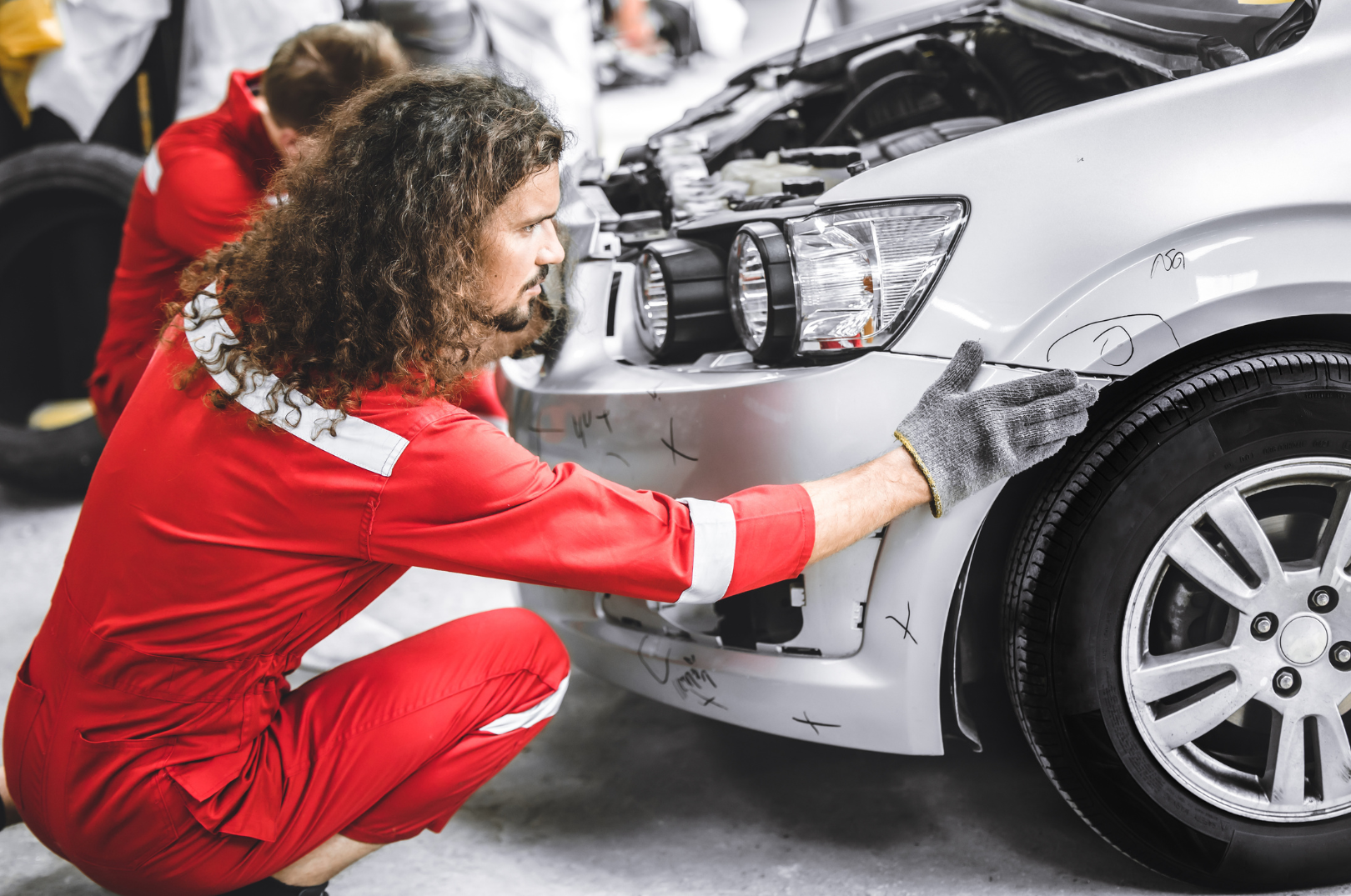 A mechanic in red coveralls inspecting the front of a silver car in a garage.