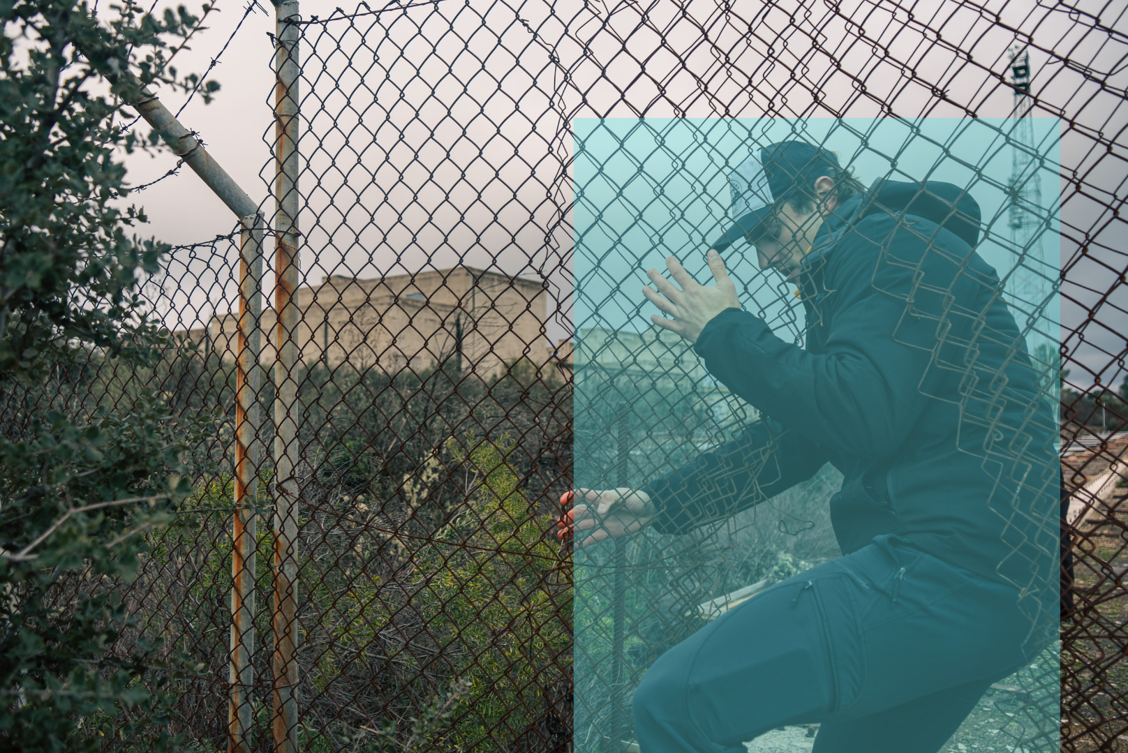 A person wearing a black jacket and cap is kneeling behind a rusted chain-link fence, attempting to break in, highlighted in a blue rectangle indicating surveillance detection, with overgrown vegetation in the background under an overcast sky.