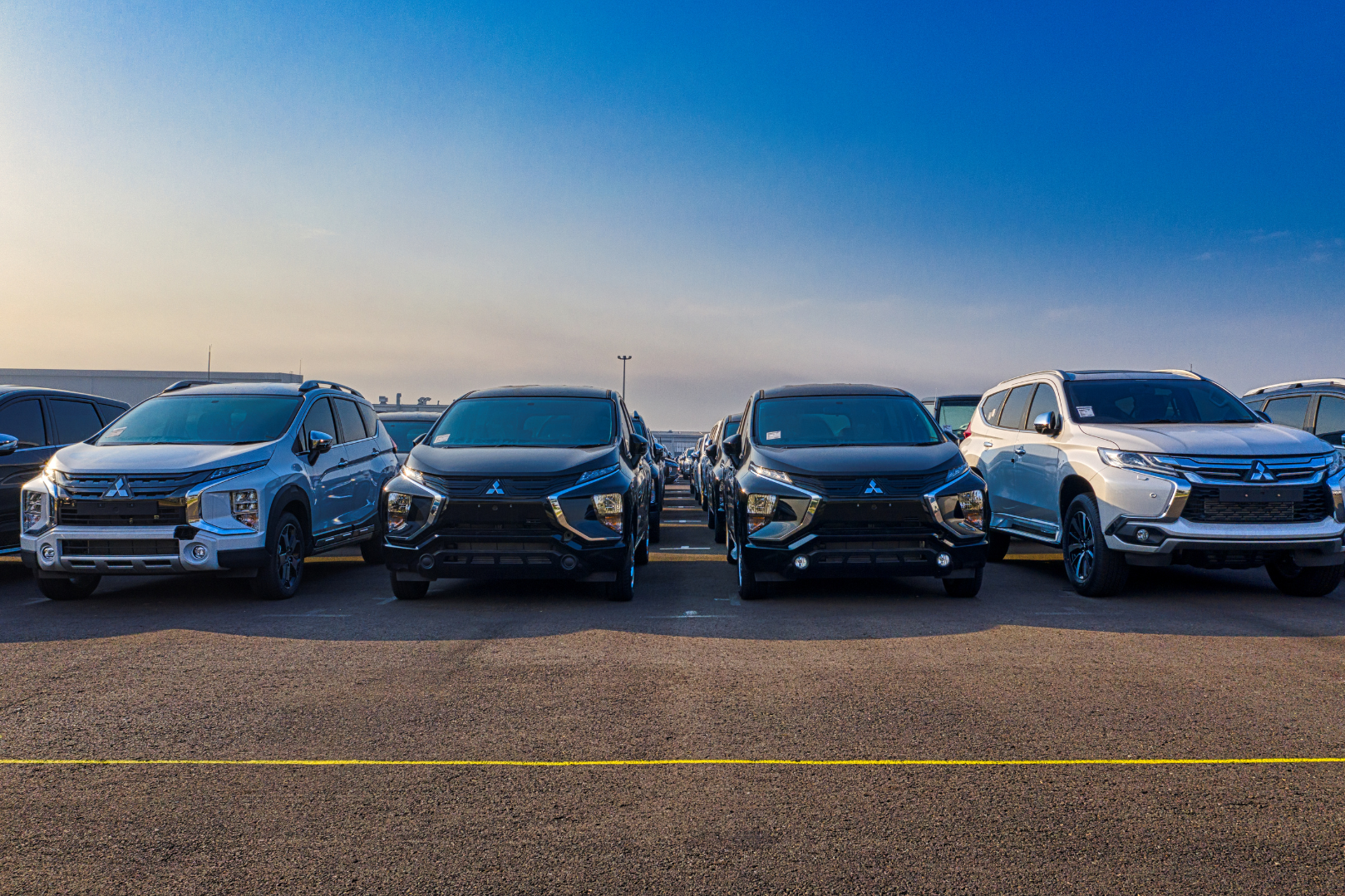 A row of four Mitsubishi SUVs parked in a parking lot under a clear blue sky, with additional cars visible in the background.