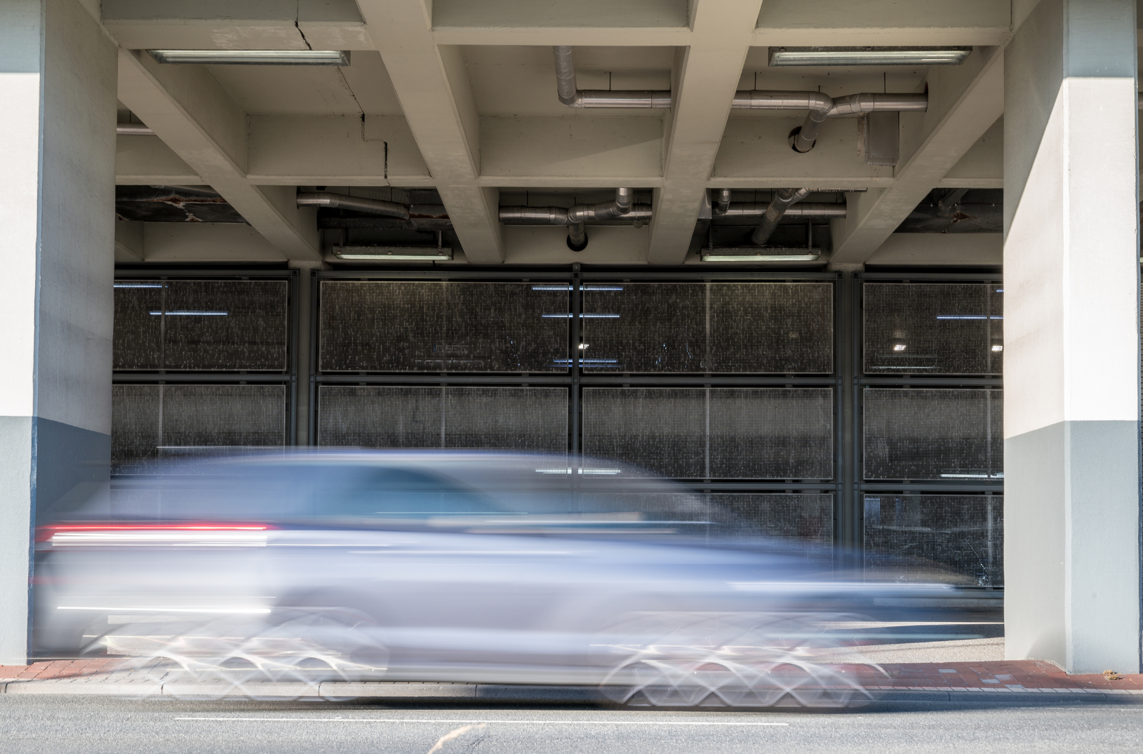 Blurred car passing under a parking structure with concrete beams and pipes, and metal fencing in the background.