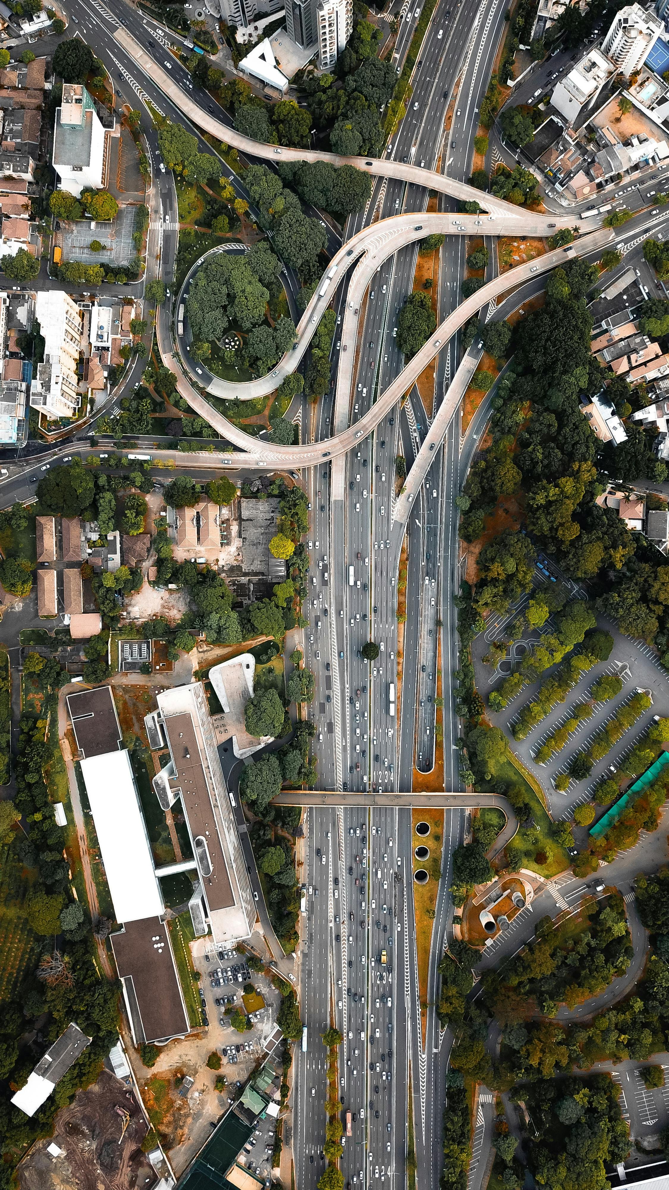 An aerial view of a busy highway with multiple lanes, overpasses, and surrounding buildings, trees, and parking lots.