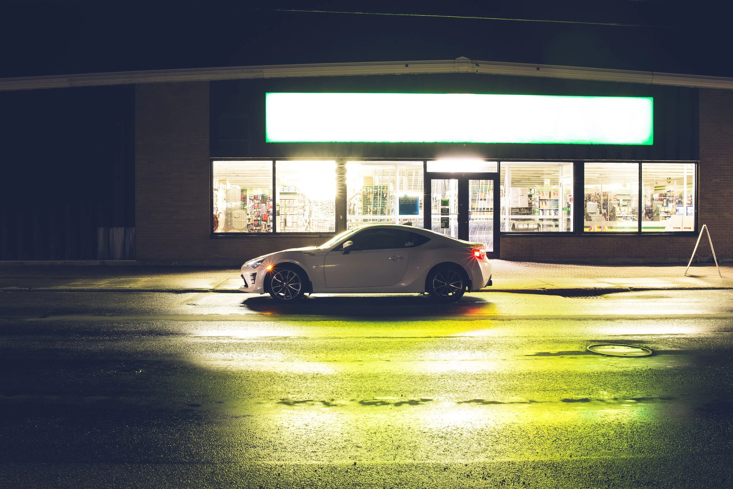 A white car parked in front of a brightly lit convenience store at night, with the store's green and white signboard visible above the entrance.