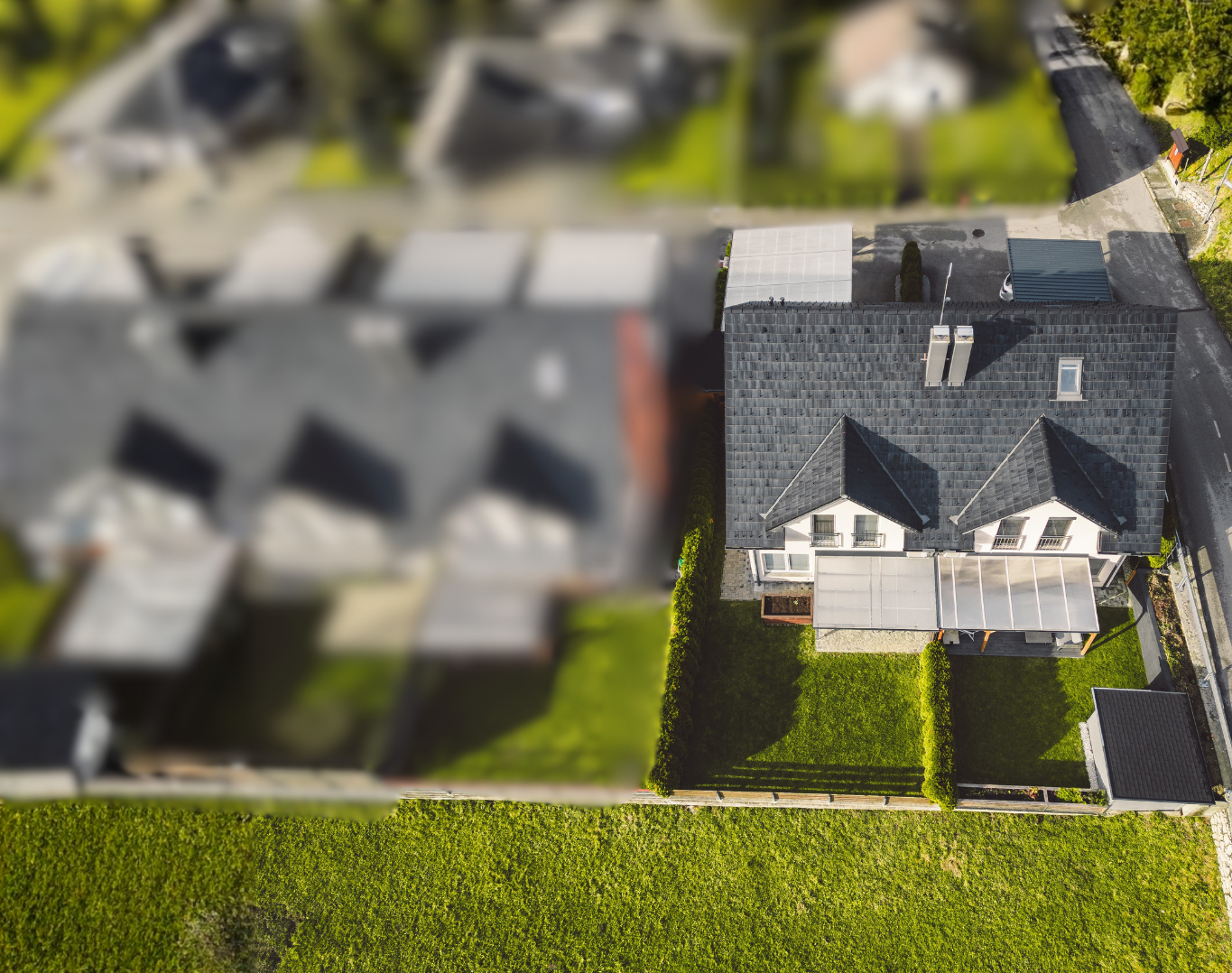 Aerial view of a house with a dark gray roof, surrounded by a green lawn, with neighboring houses and trees nearby.