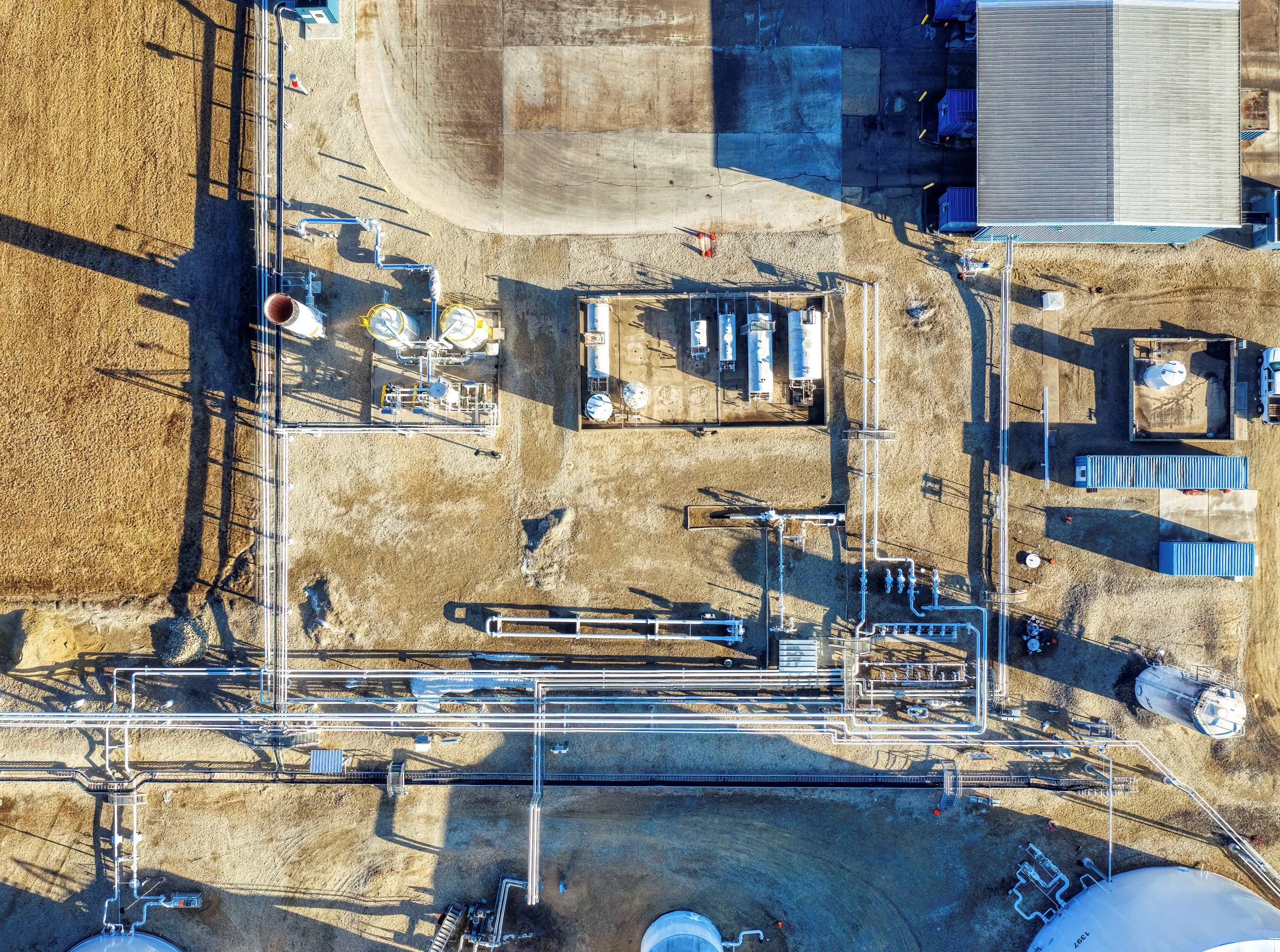 An aerial view of an industrial facility with pipelines, storage tanks, a building, and a dirt ground.