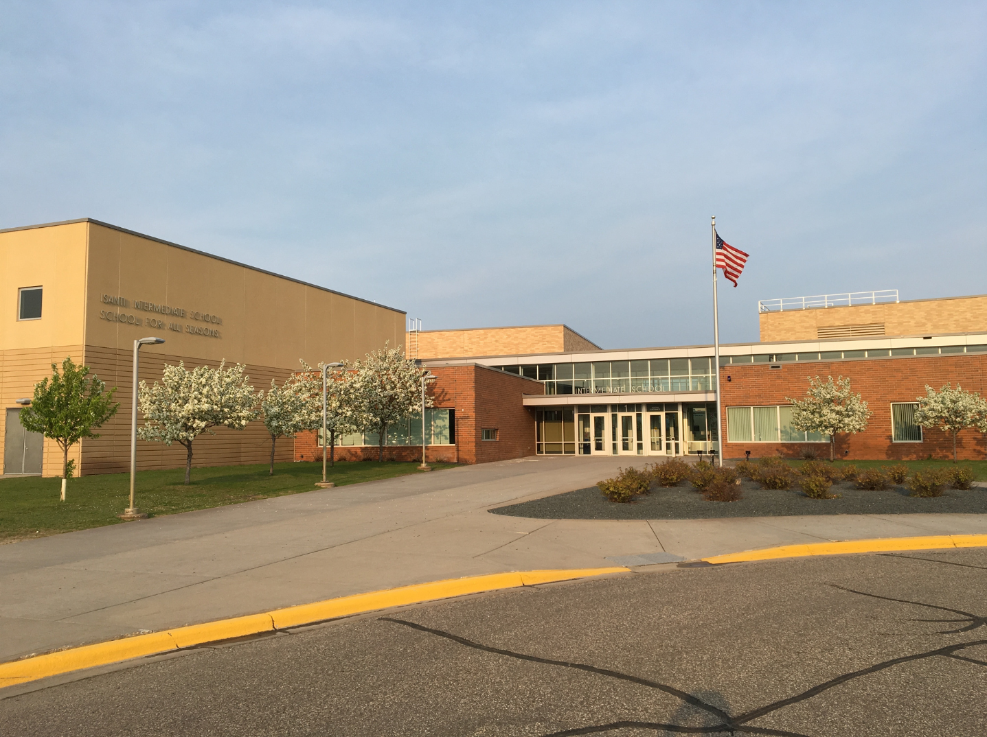 Exterior view of a school building with a sign reading 'ISANTI INTERMEDIATE SCHOOL' during sunset, with an American flag on a flagpole, some trees with blossoms, and a paved walkway leading to the entrance.