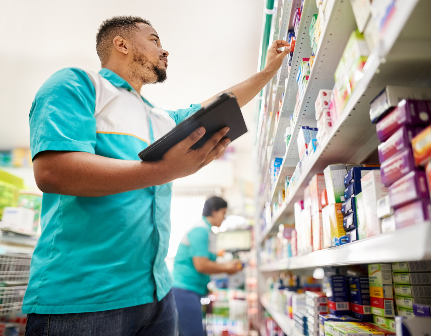 A man in a teal and white work uniform stocking medication on a store shelf, holding a black tablet and reaching for a box of medicine. Another person in similar uniform is visible in the background.