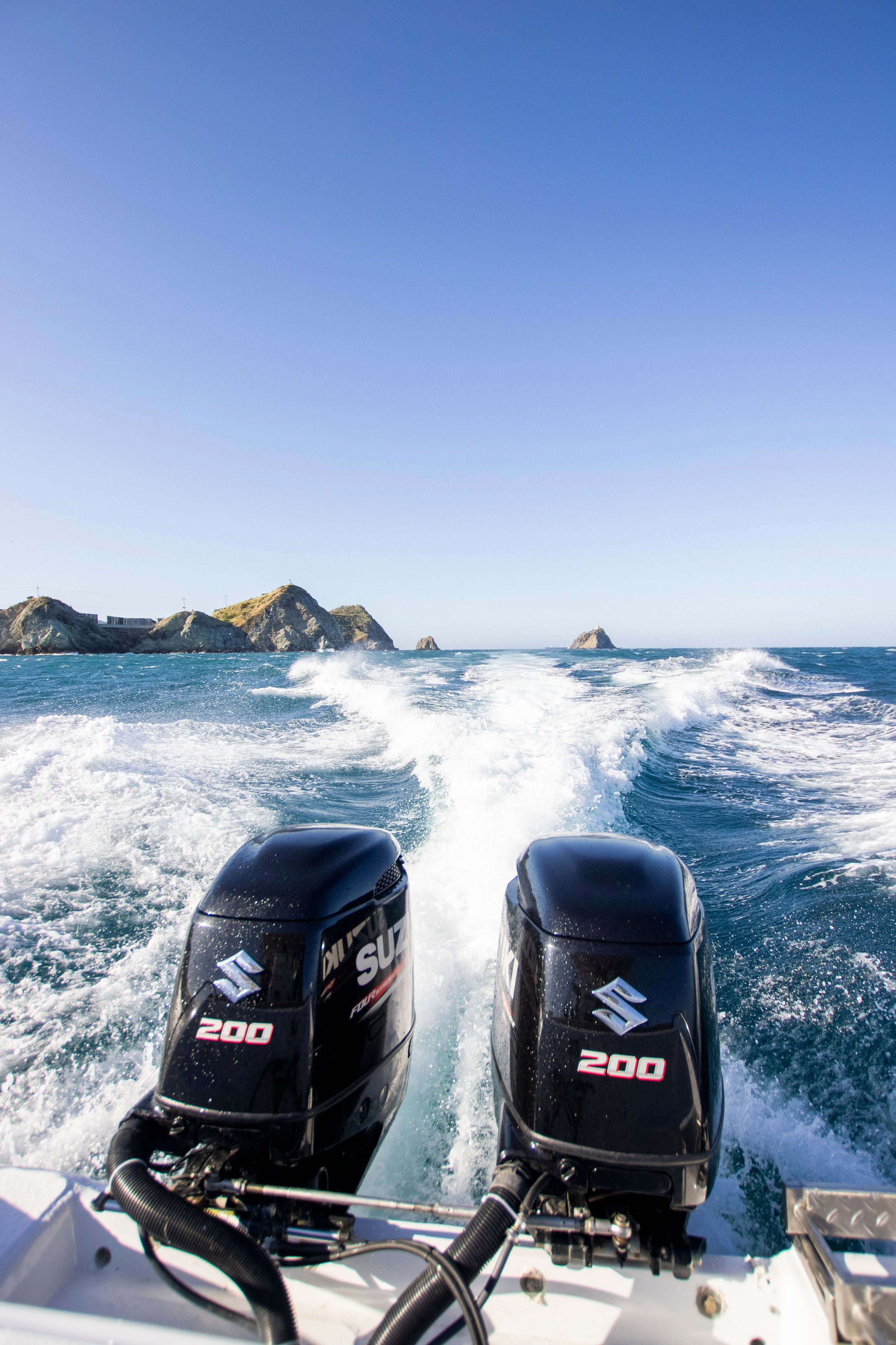 View from a boat's stern showing two Suzuki outboard motors, 200 horsepower each, propelling the boat through the ocean. Waves and a rocky island are visible in the background under a clear blue sky.