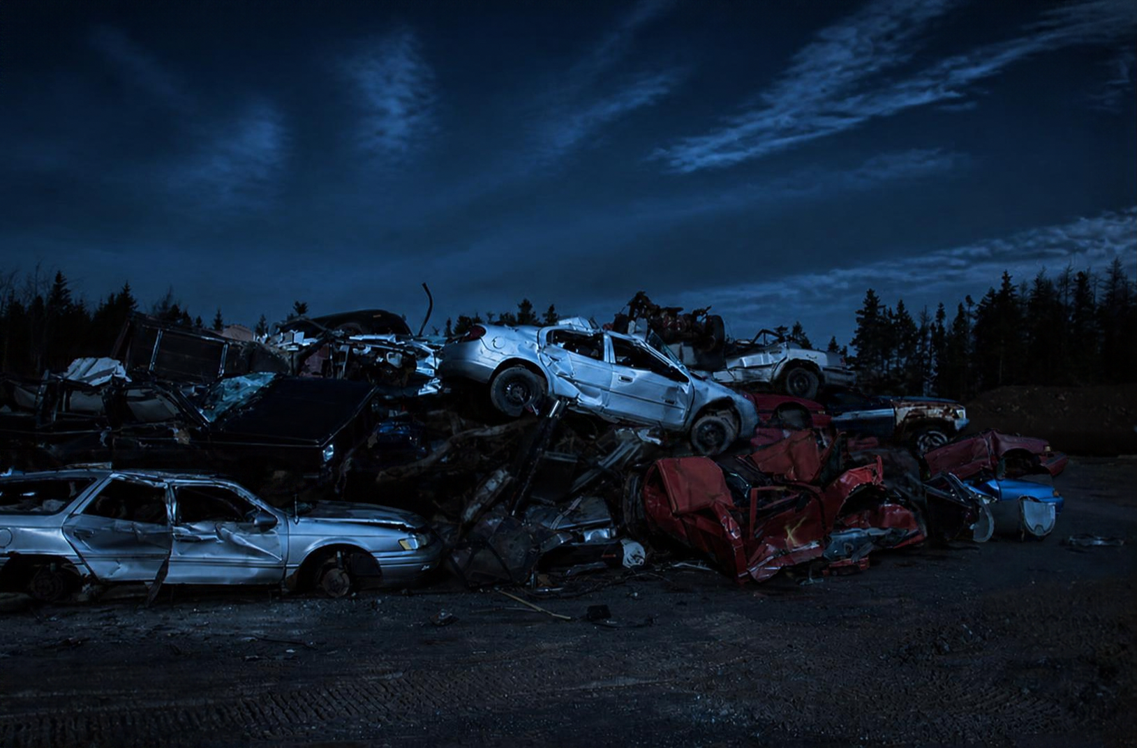 Pile of wrecked cars under a dark evening sky, with some trees in the background.