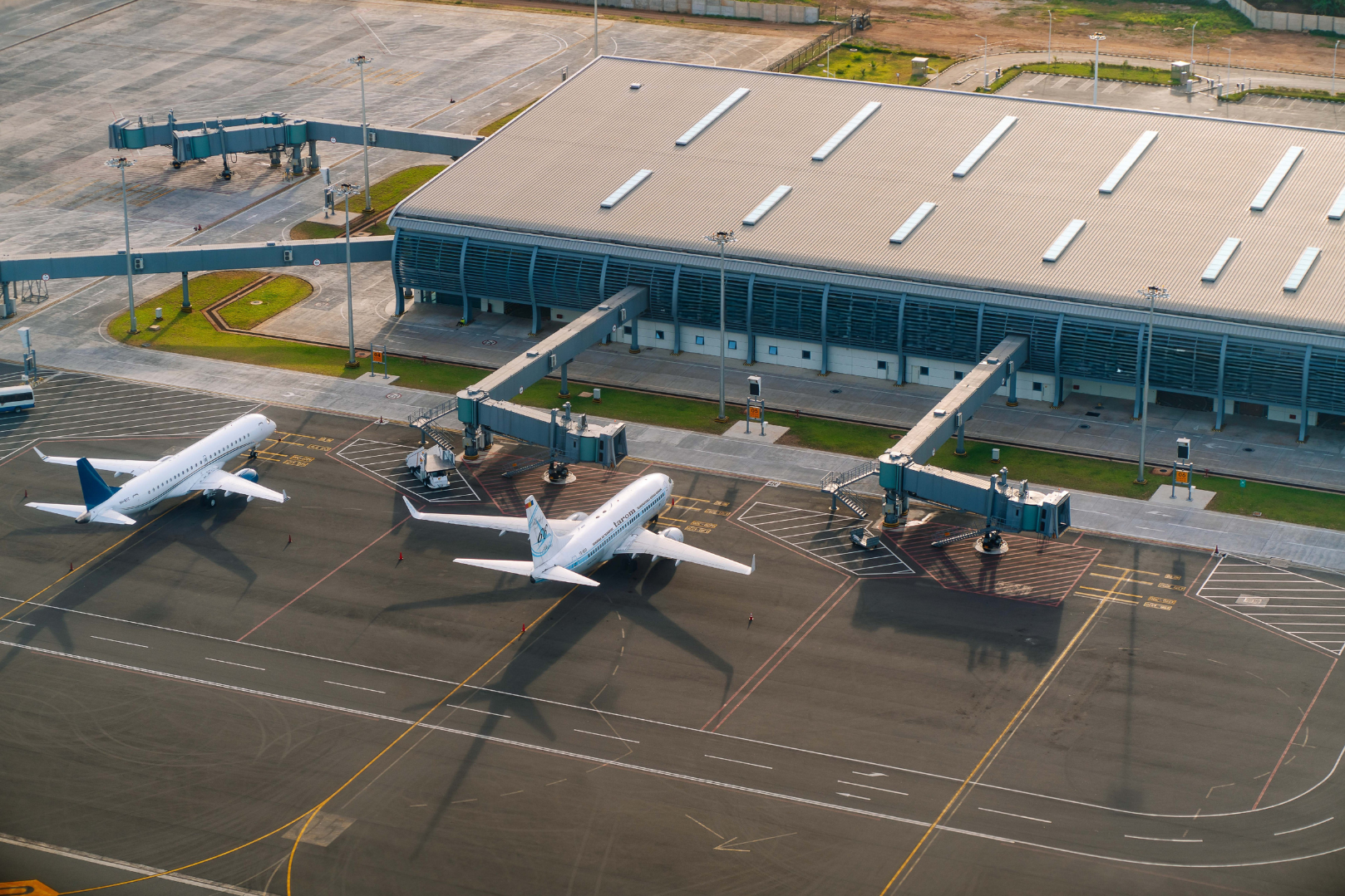 An aerial view of an airport terminal with two airplanes parked at gate, jet bridges connected, and surrounding empty tarmac and parking areas.