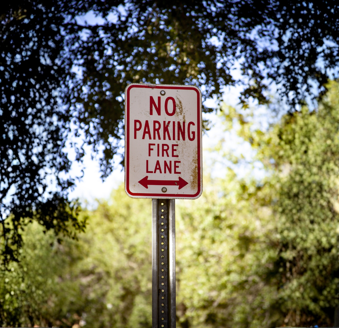 A tall sign with a white background and red text stating 'NO PARKING FIRE LANE' with arrows pointing left and right, situated outdoors among trees.
