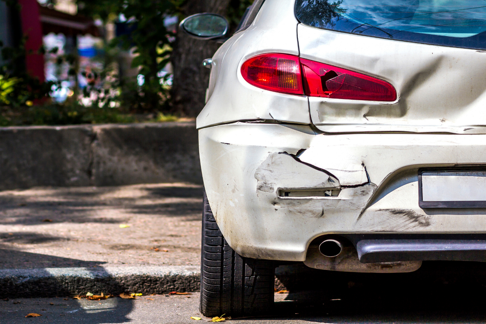 A white car with significant rear-end damage, including a cracked bumper, dented trunk, and broken tail light, parked on a street.