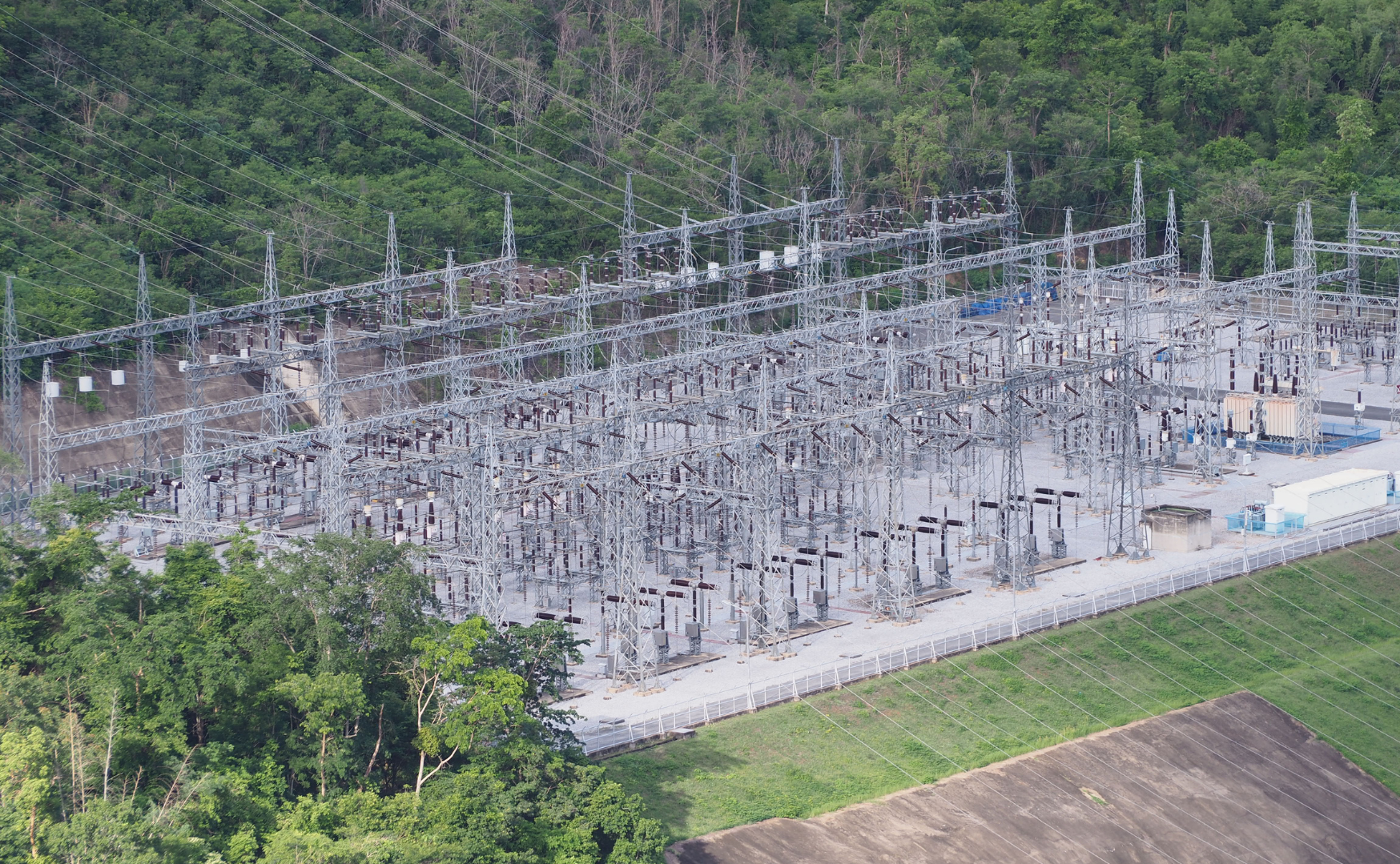 An electrical substation with numerous metal electrical towers and power lines set against a backdrop of dense green trees.