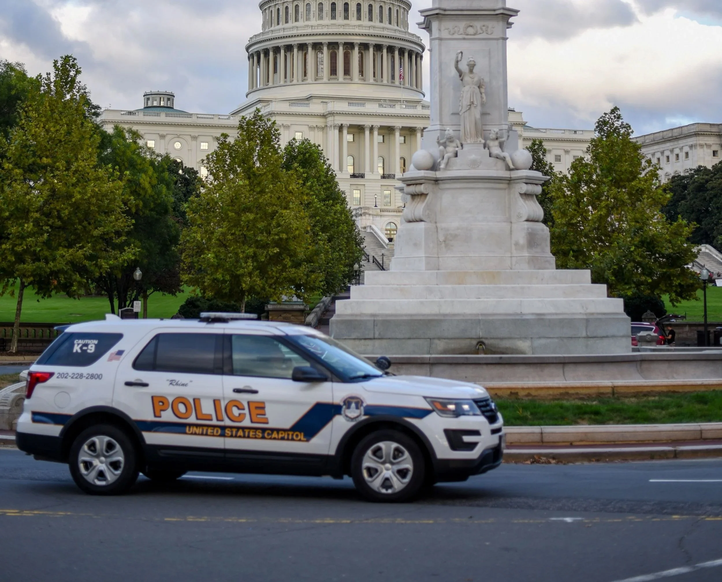 The image shows a police vehicle driving past the United States Capitol building in Washington, D.C. visible in the background, with a marble monument and trees in the scene.