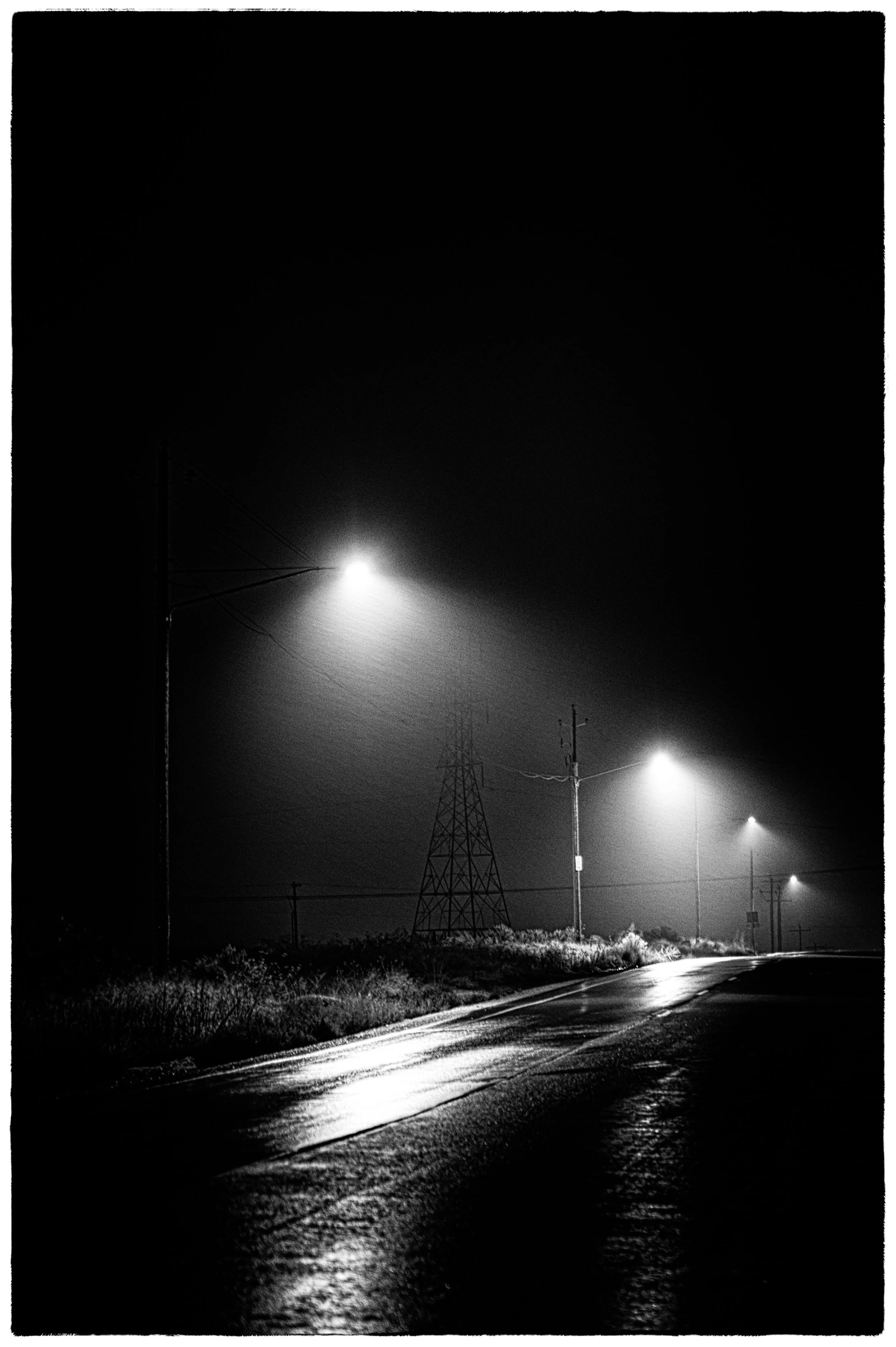 A black-and-white photo of a dark street illuminated by streetlights at night, with visible power lines, utility poles, and a transmission tower.