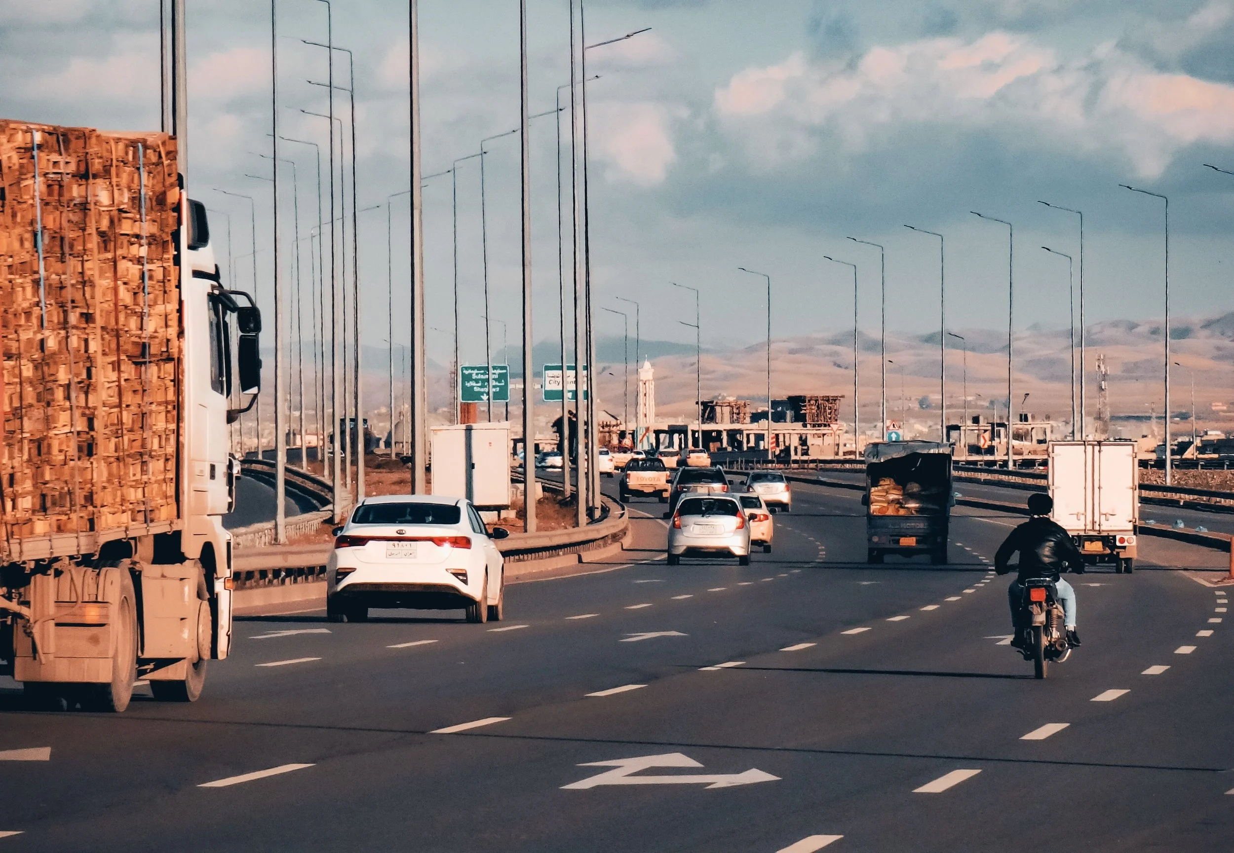 A busy highway with cars, trucks, and motorcycles, with lampposts lining the road, mountains in the background, and a partly cloudy sky.
