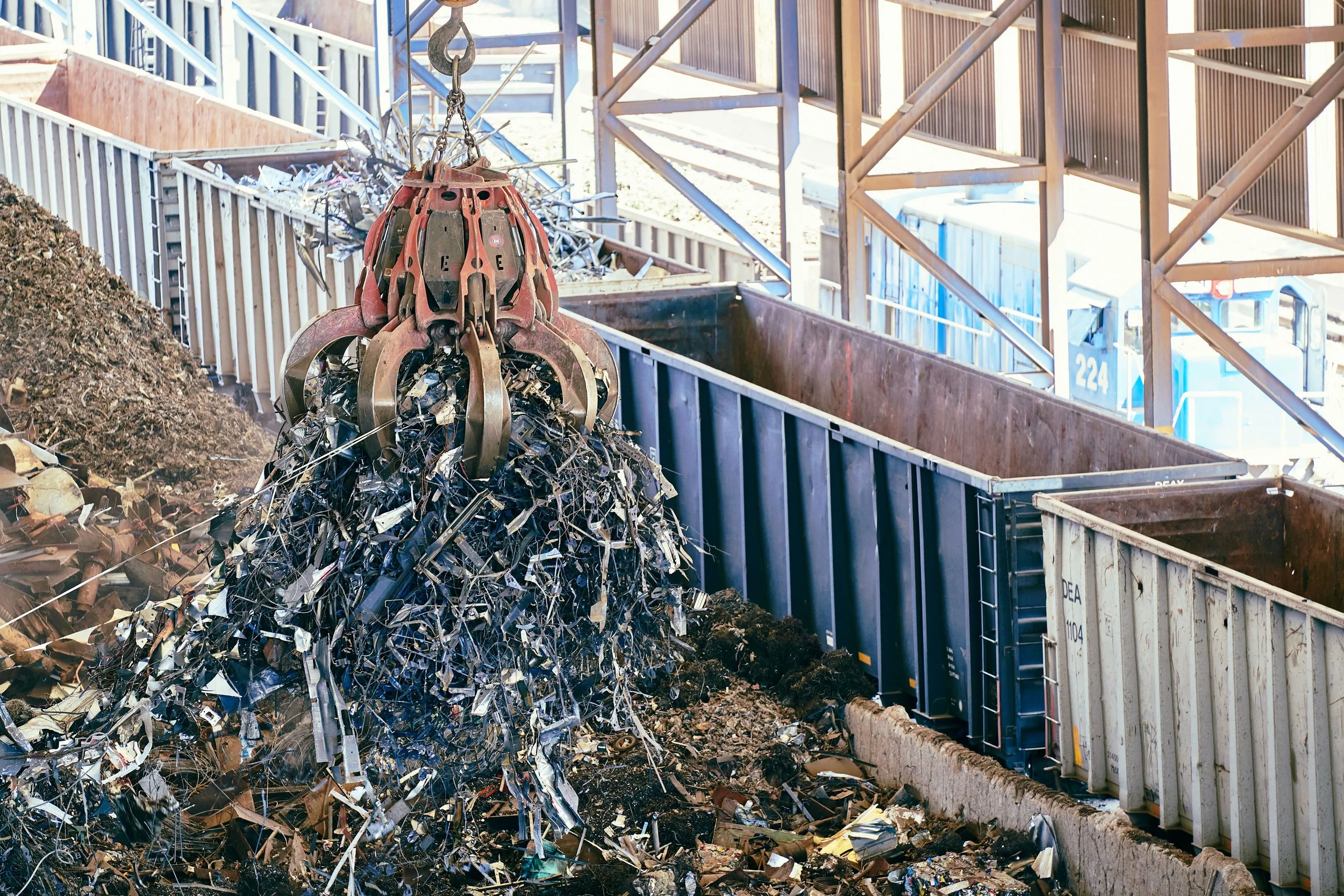 Scrap metal being loaded into a recycling bin at a recycling facility, with large mechanical claws and conveyor equipment visible in the background.