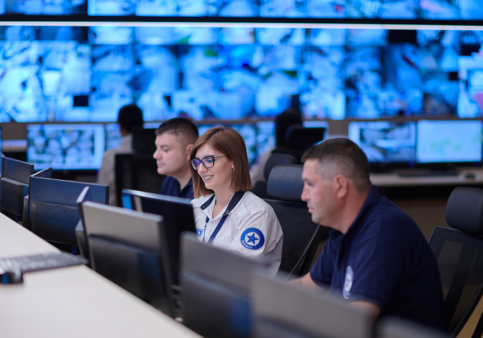 Security personnel working at computers in a security control room with multiple monitors displaying security feeds.