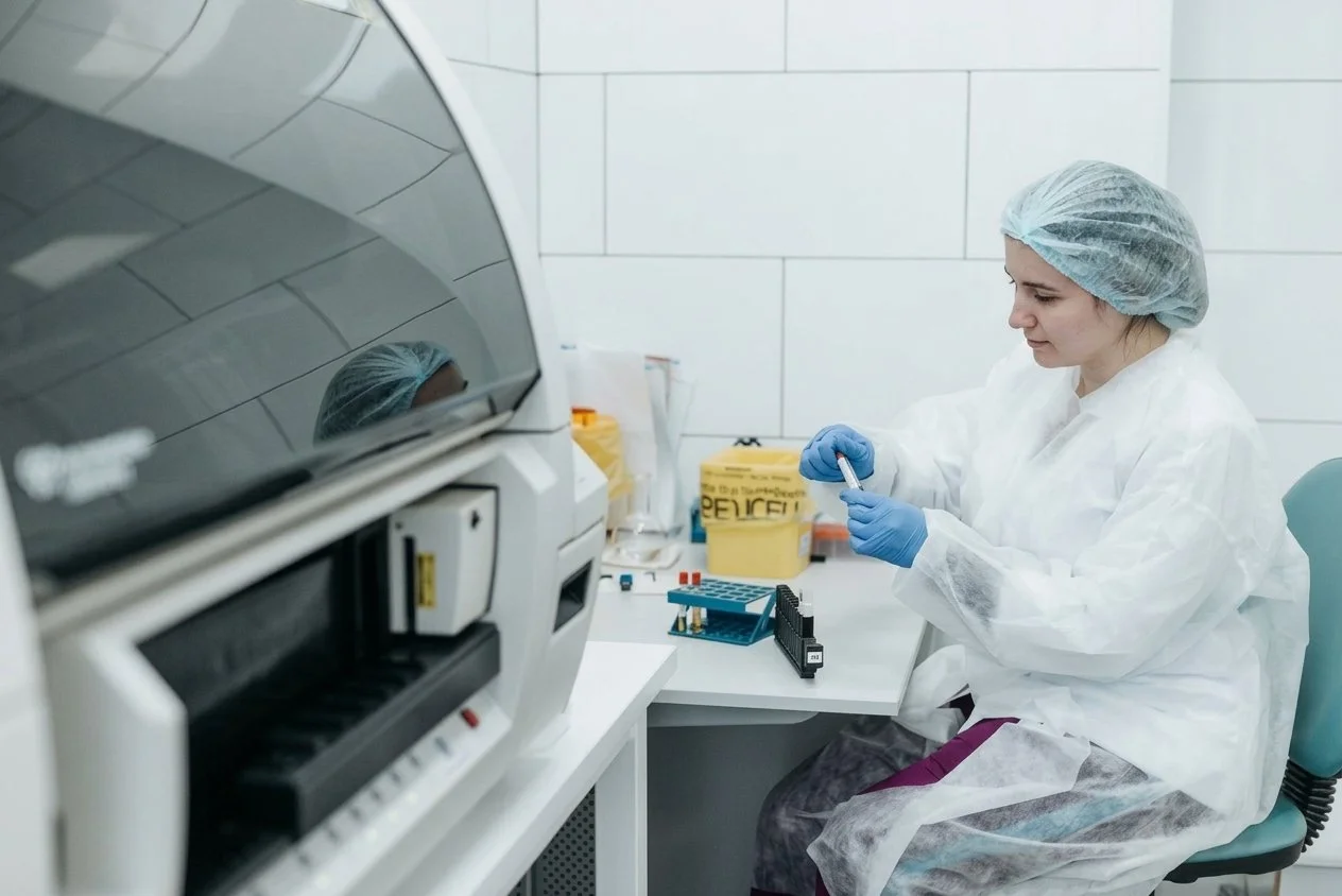 A female scientist wearing protective gear, including gloves, a lab coat, and a hair cover, working with laboratory equipment in a clean, white laboratory.
