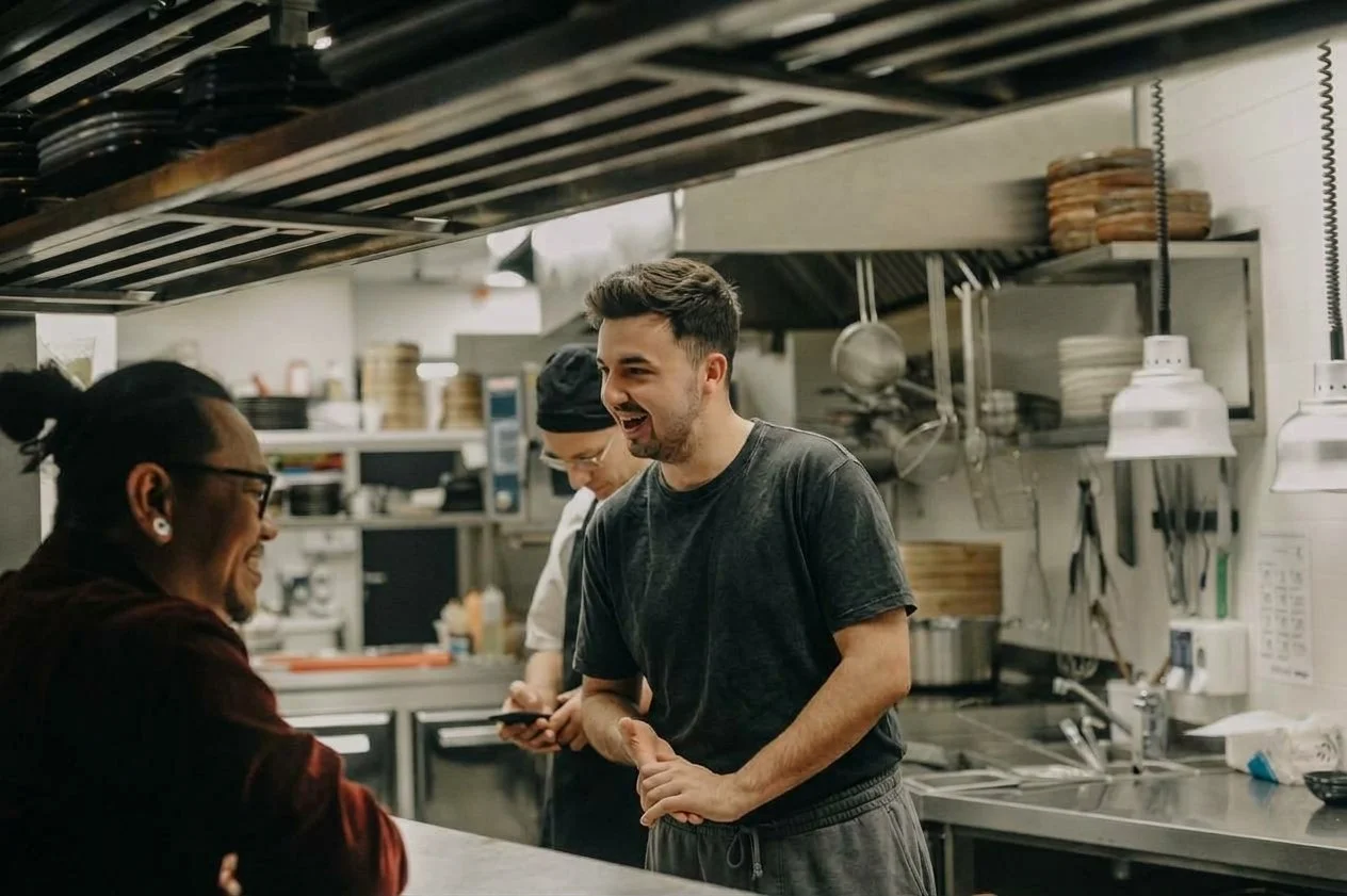 Three people laughing and talking in a commercial kitchen with stainless steel surfaces and hanging kitchen utensils.