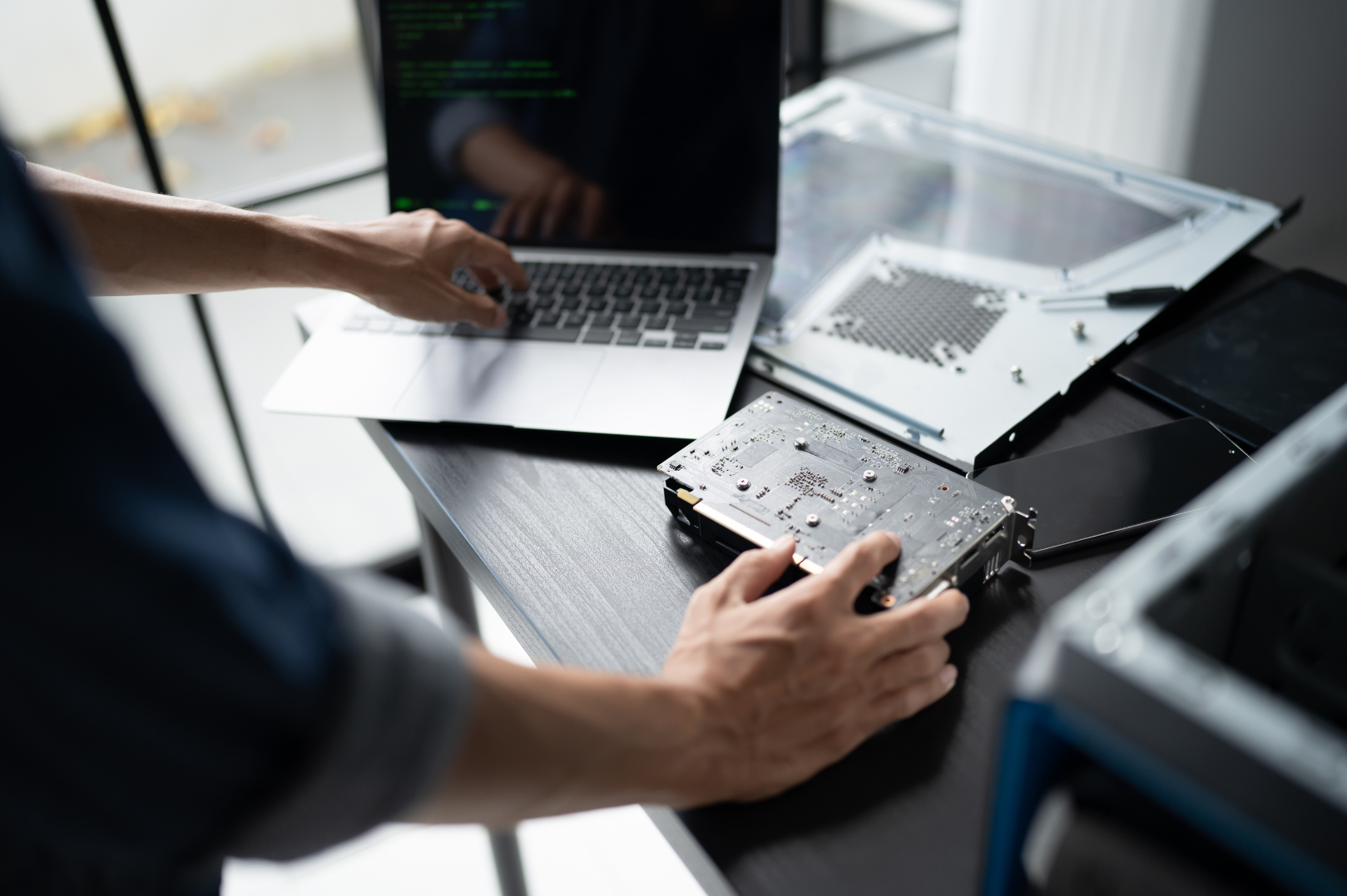 Person repairing or assembling a computer hard drive on a work desk with a laptop and electronic components surrounding it.