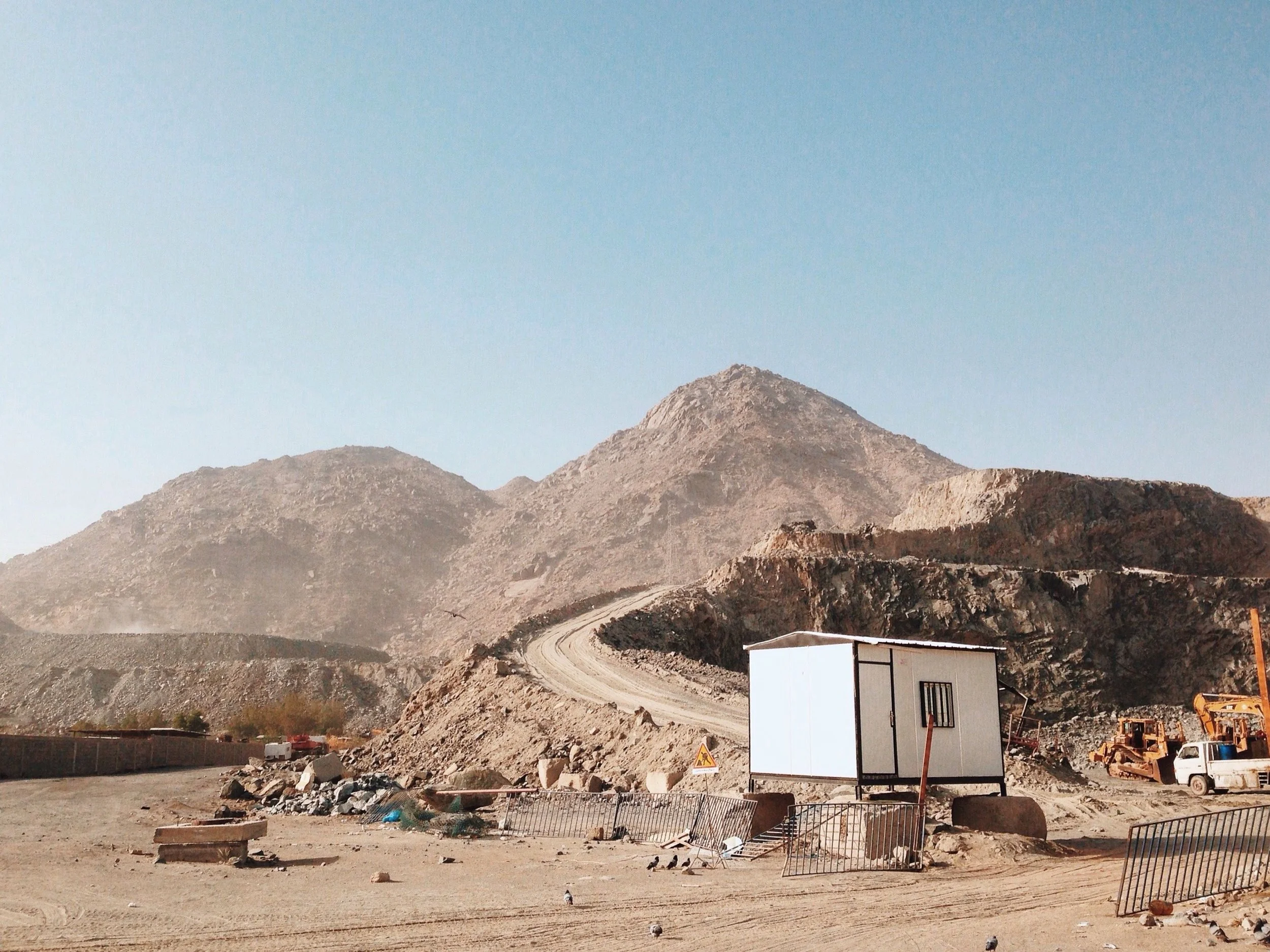 Mountain landscape with construction site in the foreground, dirt road winding up the mountain, and construction materials including small robots and a portable building.