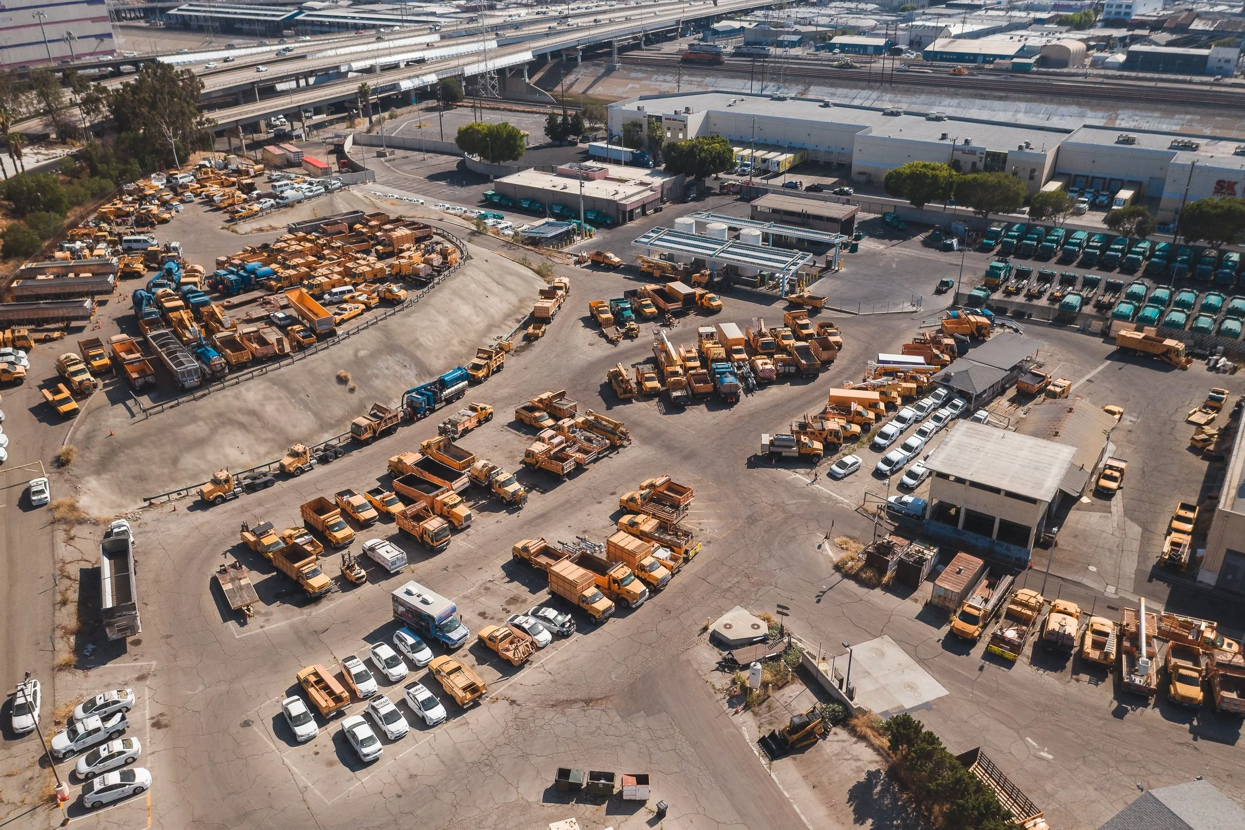 Aerial view of a parking lot with rows of silver and white cars, and a separate section with numerous yellow trucks. In the background, there are commercial buildings, trees, and elevated highways.