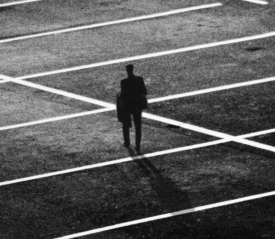 A person walking alone in an empty parking lot with marked parking spaces, casting a long shadow.