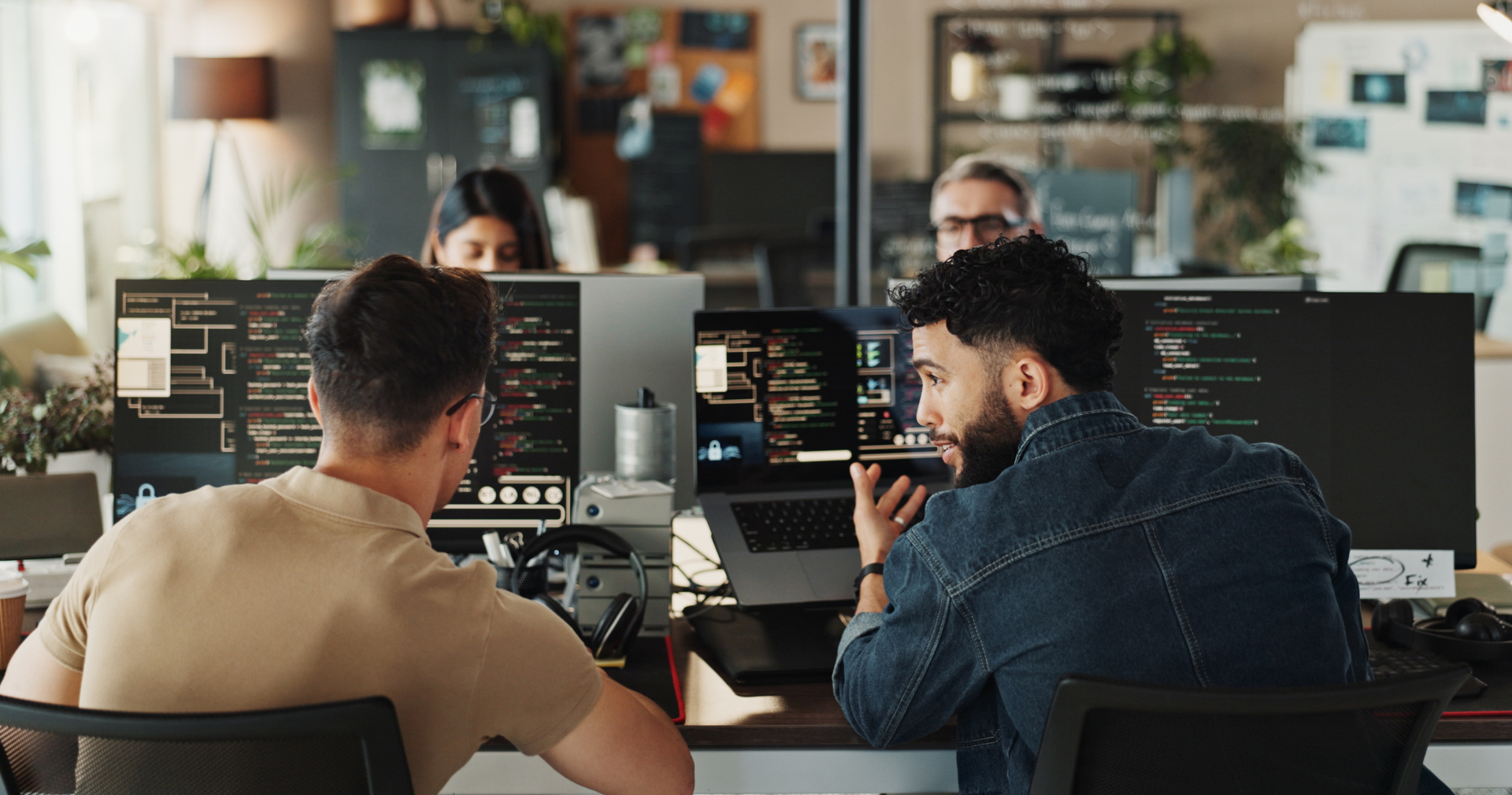 Two computer programers engaged in a conversation in a modern office, with multiple computer monitors displaying code, while other coworkers work in the background.