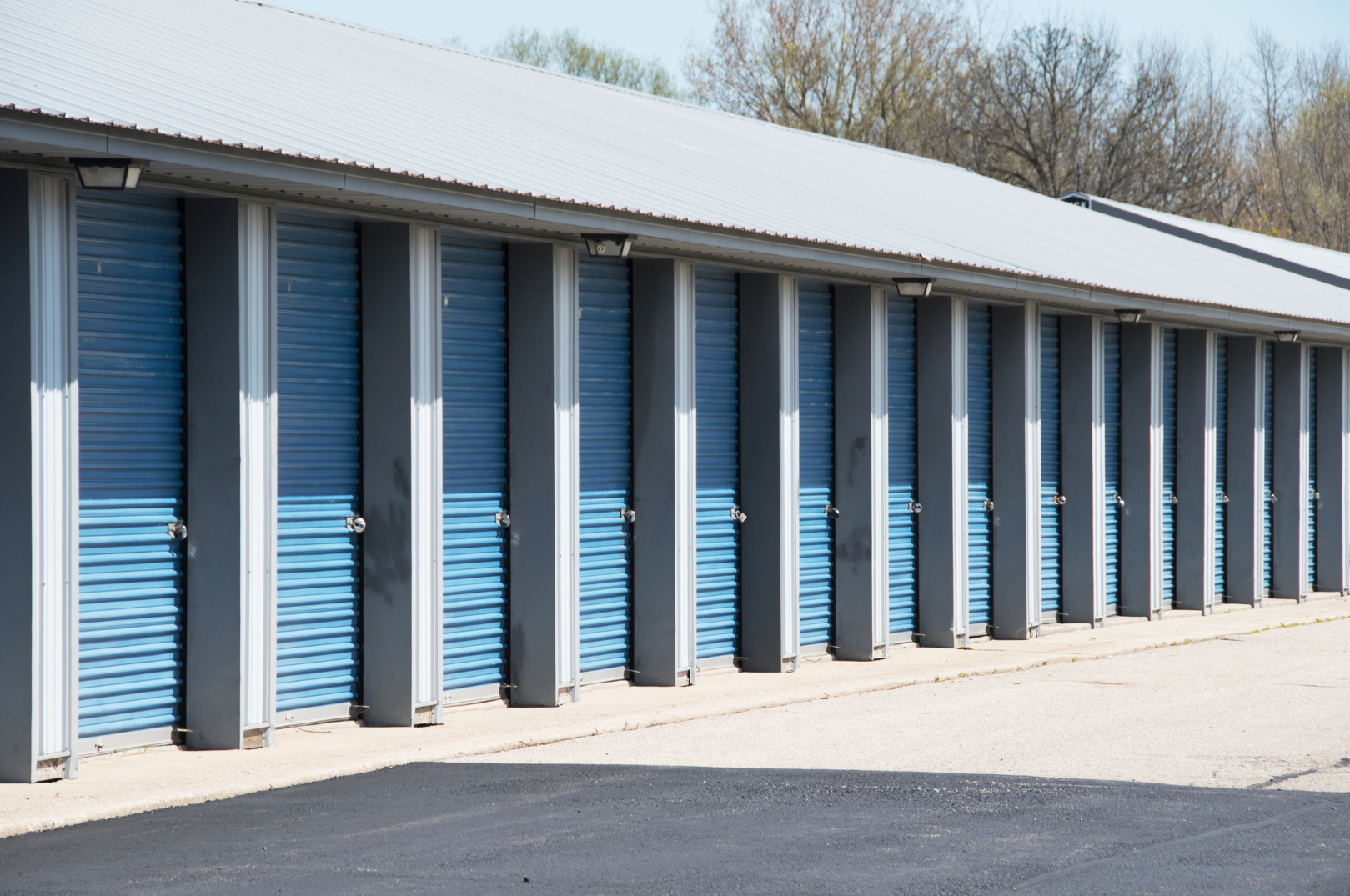 Row of blue storage units with metal roll-up doors in an outdoor storage facility, cleared paved ground, and leafless trees in the background.
