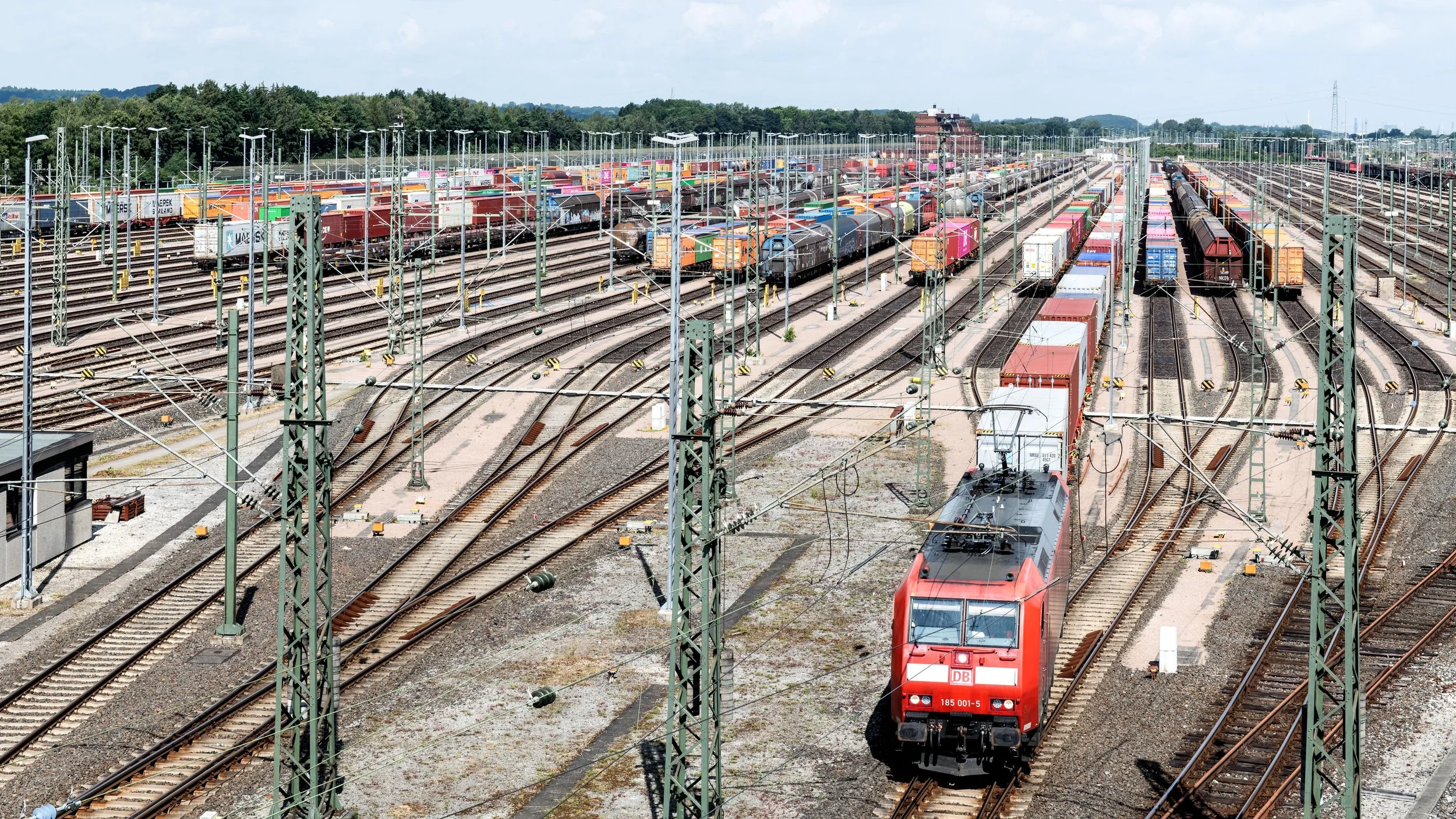 A large freight train yard with numerous train tracks and a red locomotive train in the foreground. Multiple freight cars are visible on the tracks, with a green landscape and trees in the background under a partly cloudy sky.
