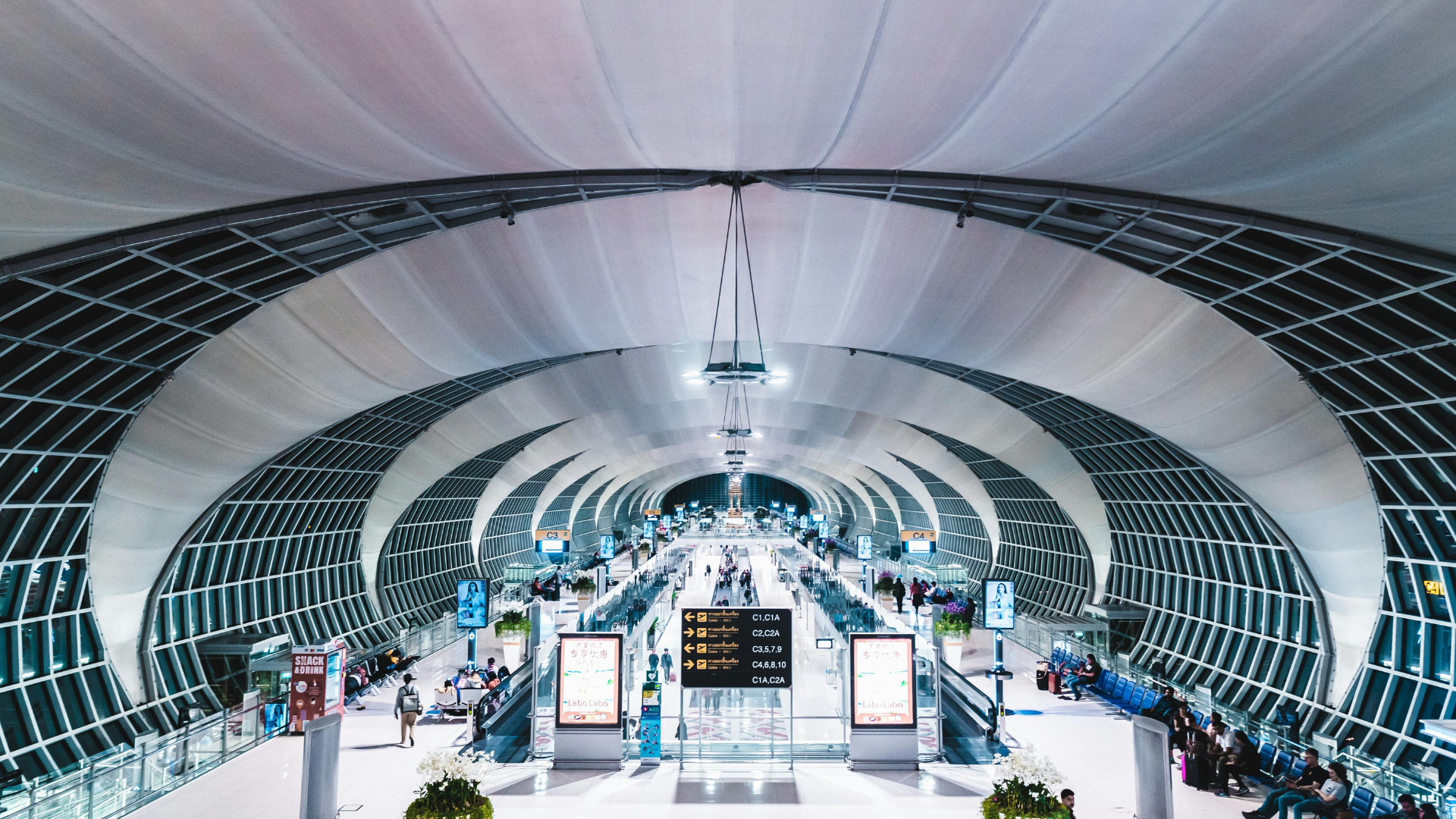 Interior view of a modern airport terminal with a curved ceiling, multiple gates, and travelers walking or sitting.