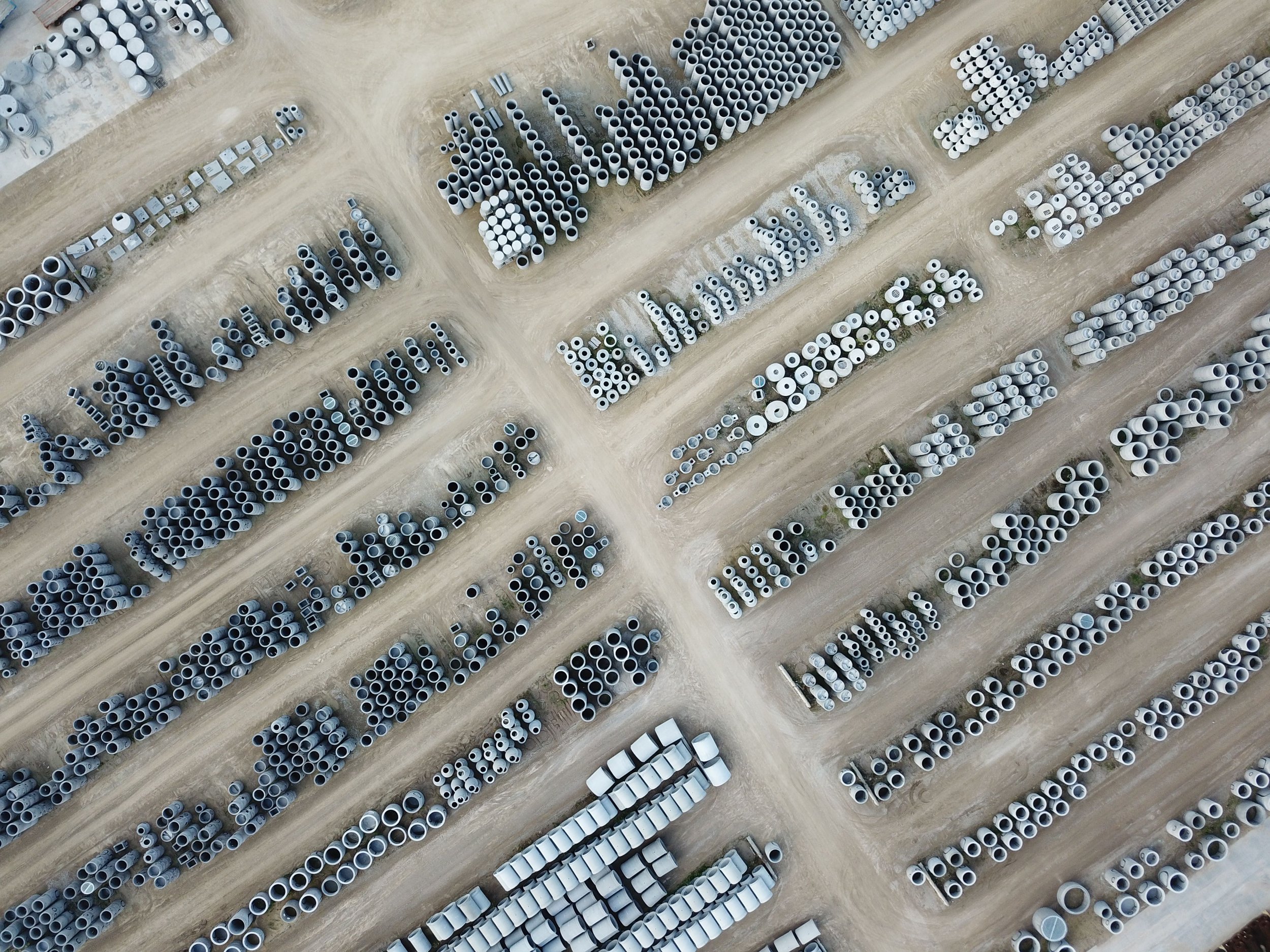 An aerial view of a large outdoor storage yard with rows of concrete pipes and other construction materials arranged on dirt ground.